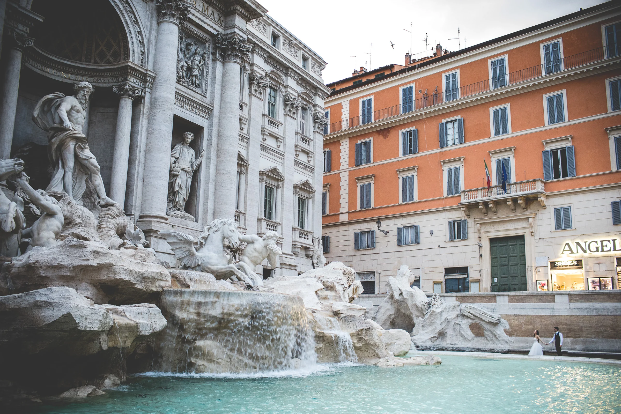 Vista amplia de la Fontana di Trevi con los novios al borde del agua durante una sesión postboda en Fontana di Trevi.
