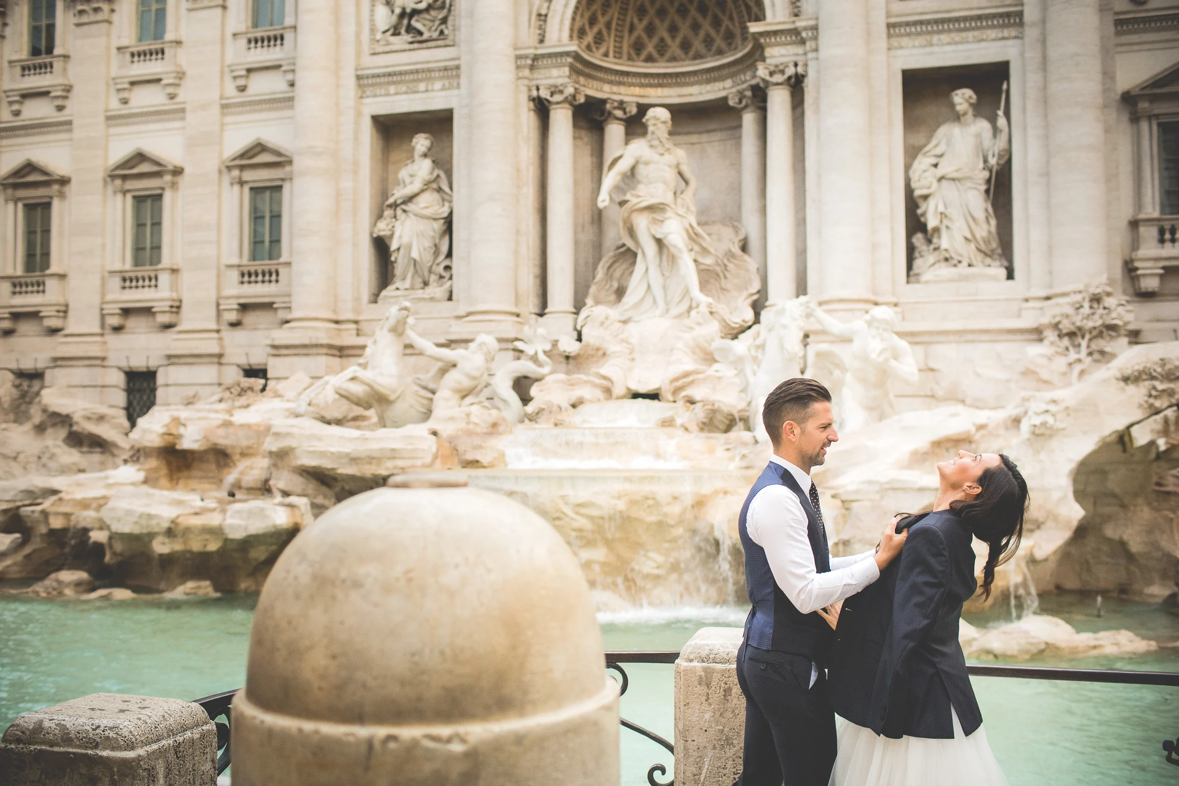 El novio ayuda a la novia a ponerse una chaqueta frente a la Fontana di Trevi durante una sesión postboda en Fontana di Trevi.