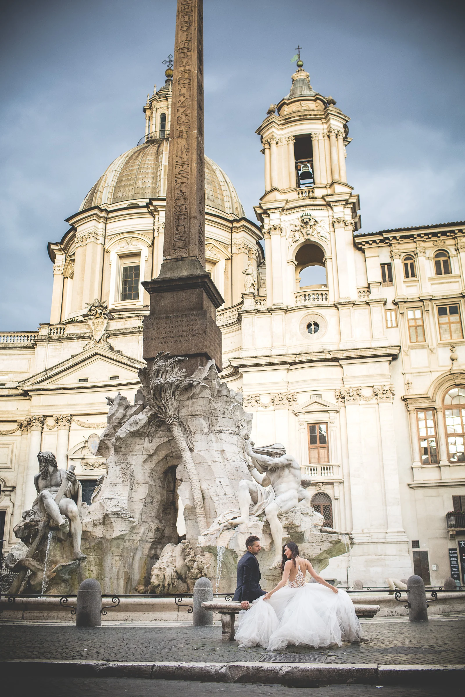 La pareja sentada junto a la Fontana dei Quattro Fiumi y el obelisco de Piazza Navona durante una sesión postboda en Piazza Navona.