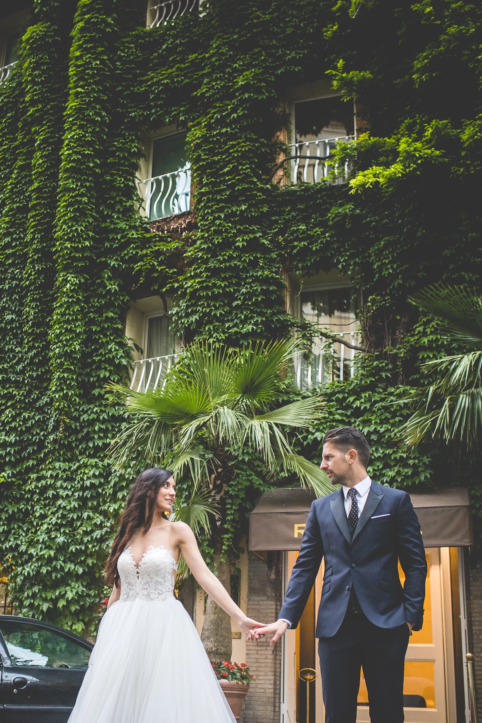 Los novios caminan de la mano ante una fachada cubierta de hiedra durante una sesión postboda en Centro histórico de Roma.