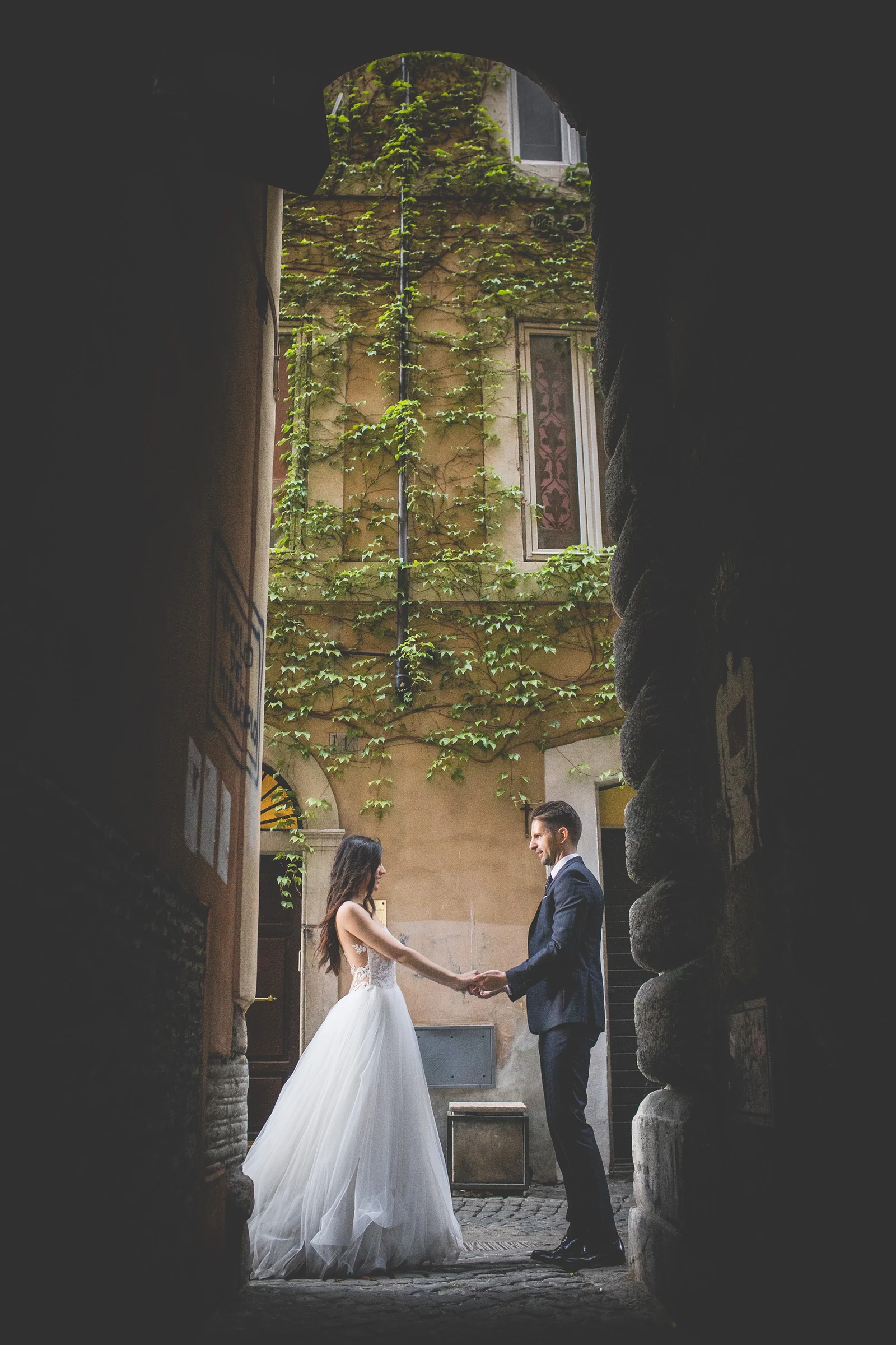 Los novios se miran bajo un arco oscuro con una fachada cubierta de hiedra al fondo durante una sesión postboda en Centro histórico de Roma.