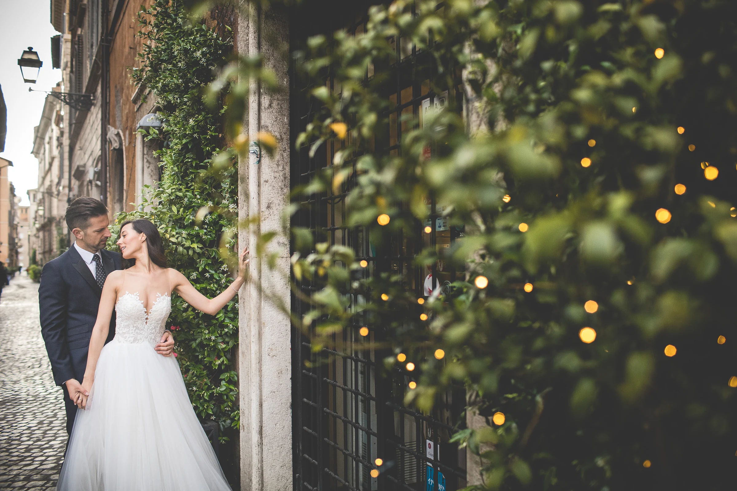 Los novios posan junto a una entrada rodeada de plantas y pequeñas luces durante una sesión postboda en Centro histórico de Roma.