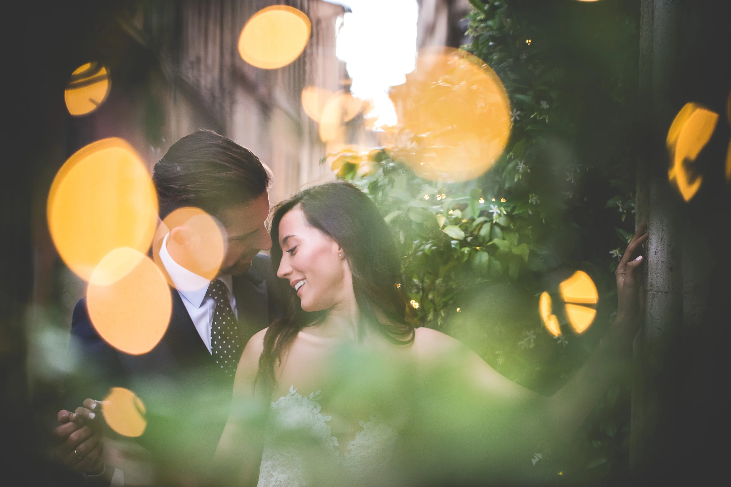 Retrato creativo de la pareja entre hojas desenfocadas y luces cálidas de bokeh durante una sesión postboda en Centro histórico de Roma.
