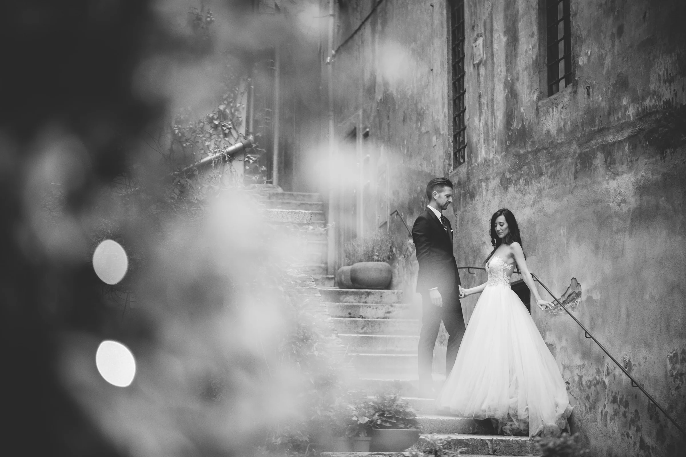 Retrato en blanco y negro de la pareja en una escalinata con luces desenfocadas en primer plano durante una sesión postboda en Centro histórico de Roma.