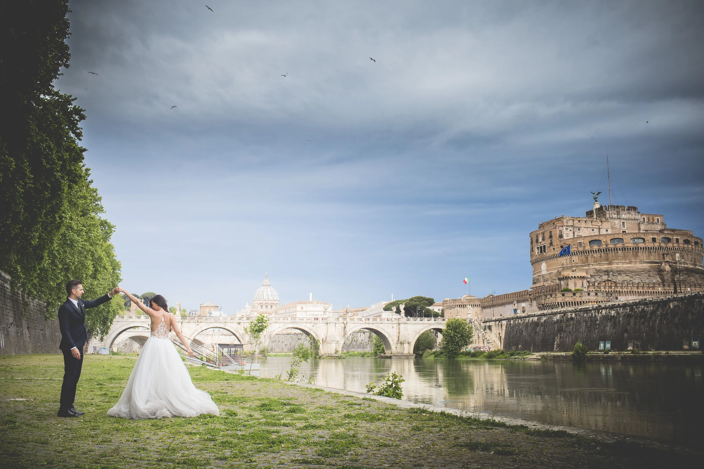 Vista panorámica de la pareja junto al Tíber con Castel Sant’Angelo y San Pedro al fondo durante una sesión postboda en Ponte Sant’Angelo y río Tíber.
