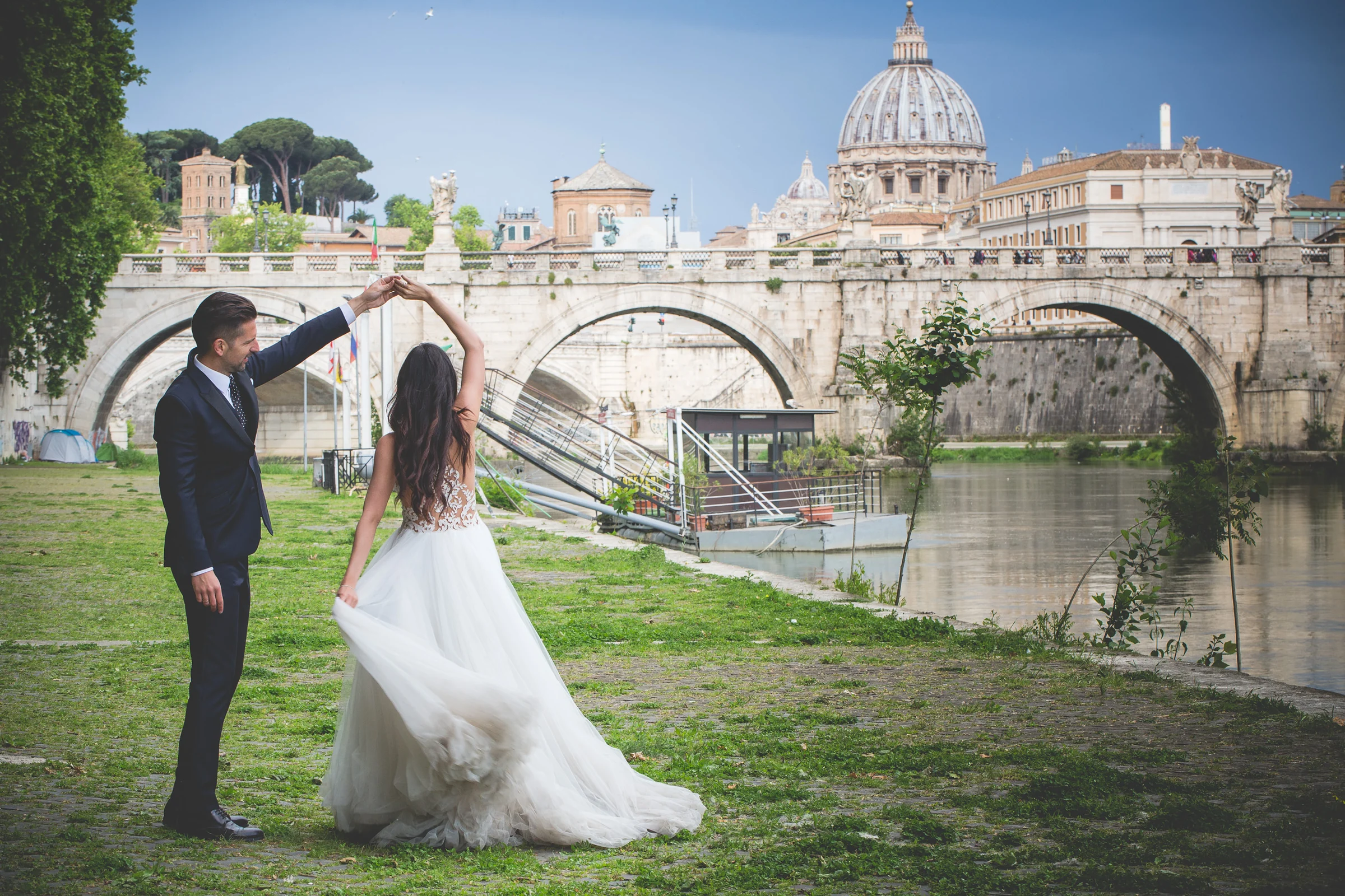 Los novios bailan junto al Tíber con la cúpula de San Pedro al fondo durante una sesión postboda en Ponte Sant’Angelo y río Tíber.