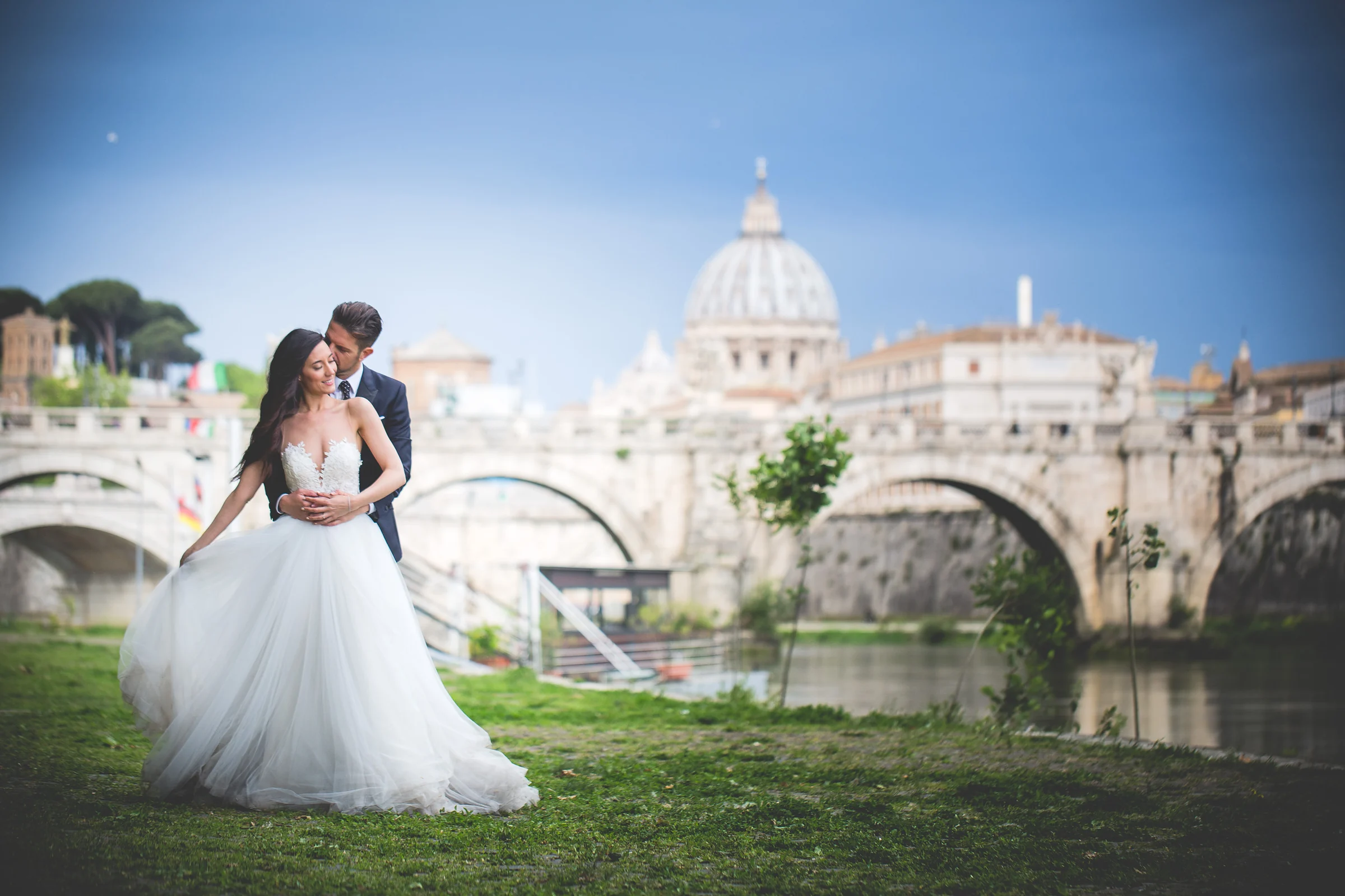 La pareja abrazada junto al río con el puente y la Basílica de San Pedro de fondo durante una sesión postboda en Ponte Sant’Angelo y río Tíber.