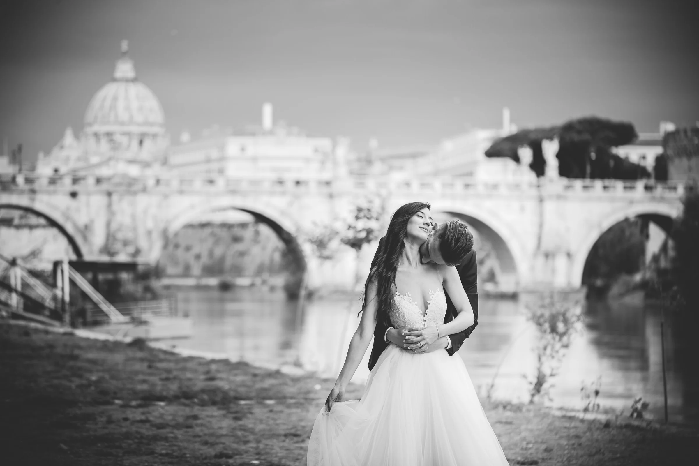Retrato en blanco y negro de los novios junto al Tíber con San Pedro al fondo durante una sesión postboda en Ponte Sant’Angelo y río Tíber.