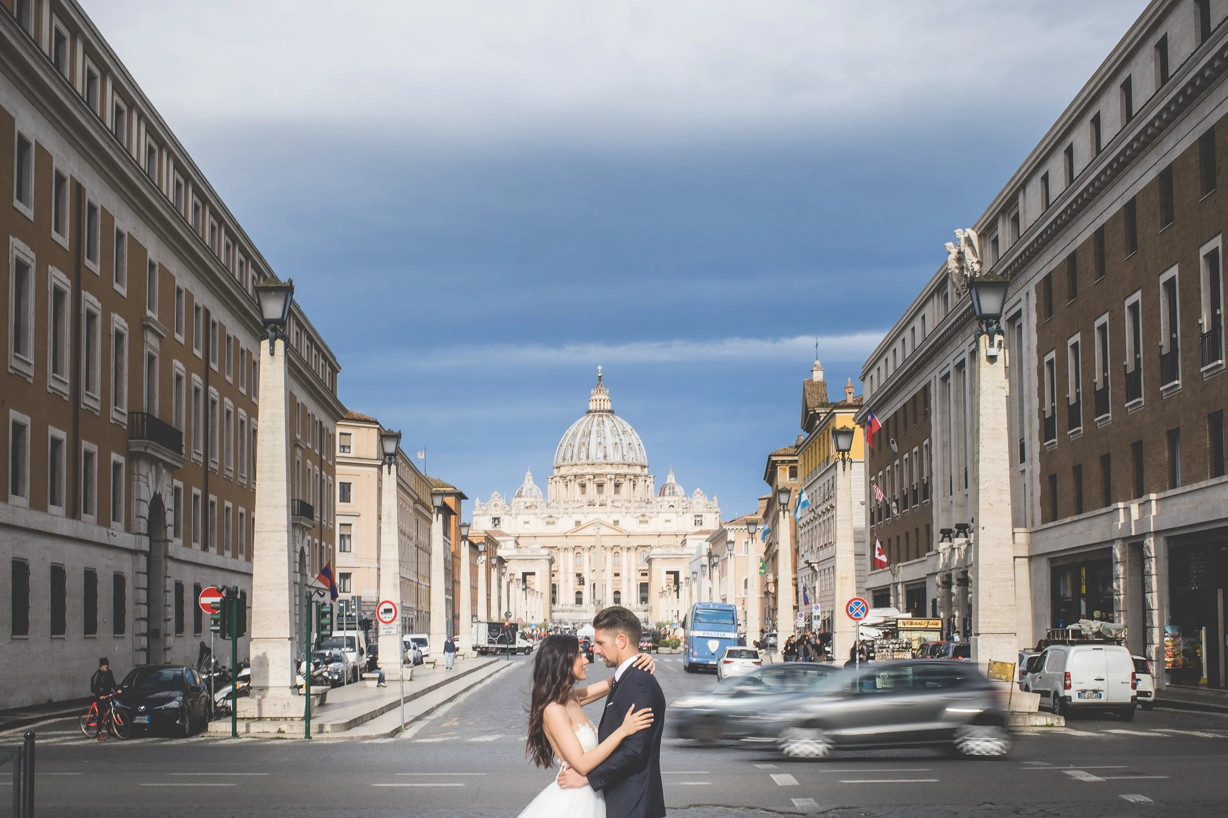 Los novios se abrazan en una gran avenida con la Basílica de San Pedro al fondo y tráfico en movimiento durante una sesión postboda en Vaticano y Plaza de San Pedro.
