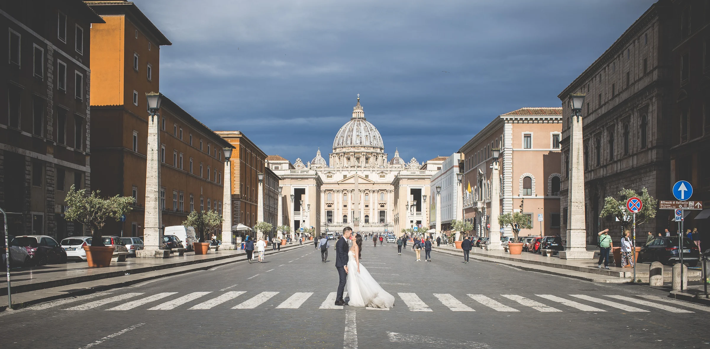 Vista simétrica de la pareja en el centro de Via della Conciliazione con San Pedro al fondo durante una sesión postboda en Vaticano y Plaza de San Pedro.