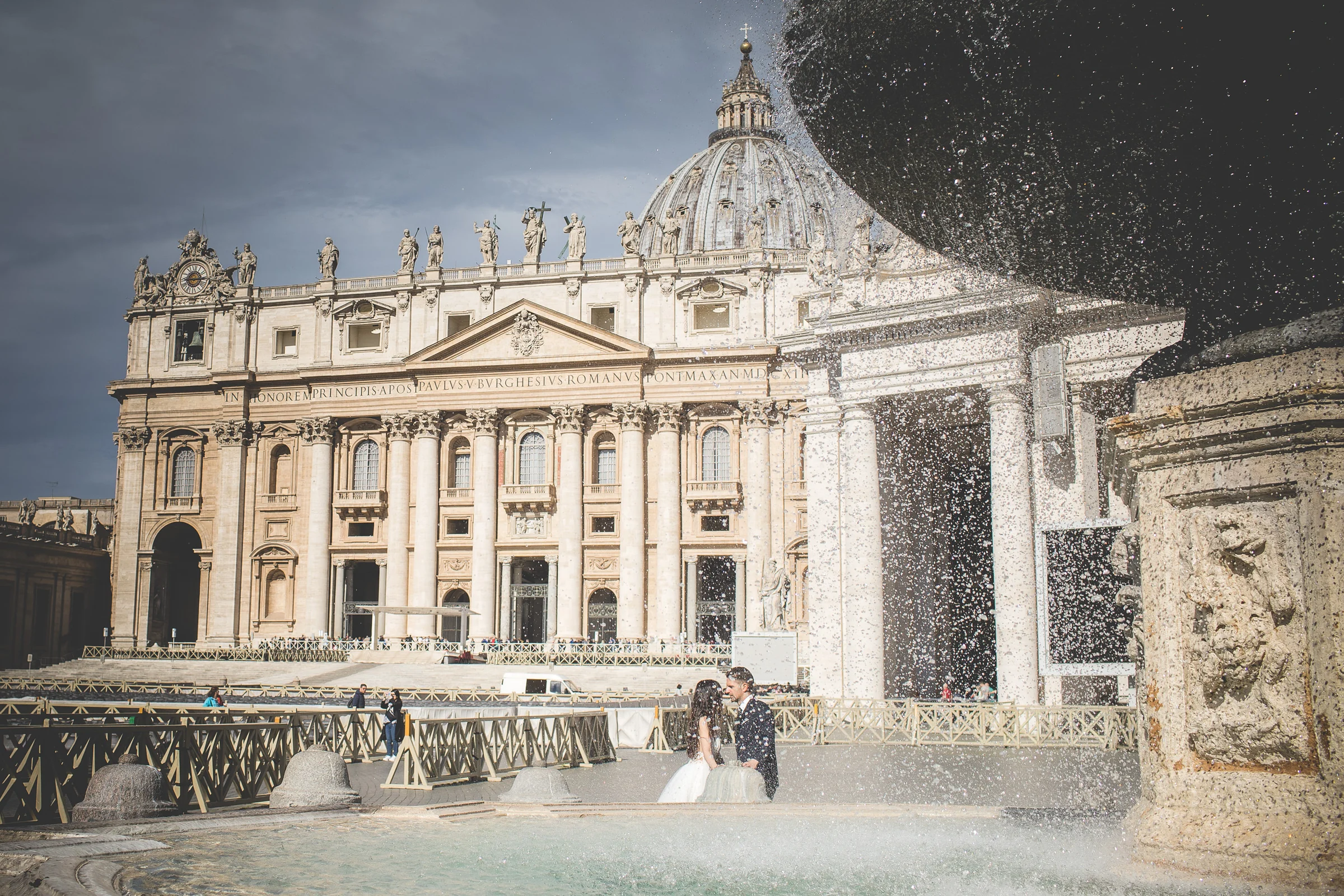 Vista amplia de la Basílica de San Pedro con la pareja junto a una fuente en primer plano durante una sesión postboda en Vaticano y Plaza de San Pedro.