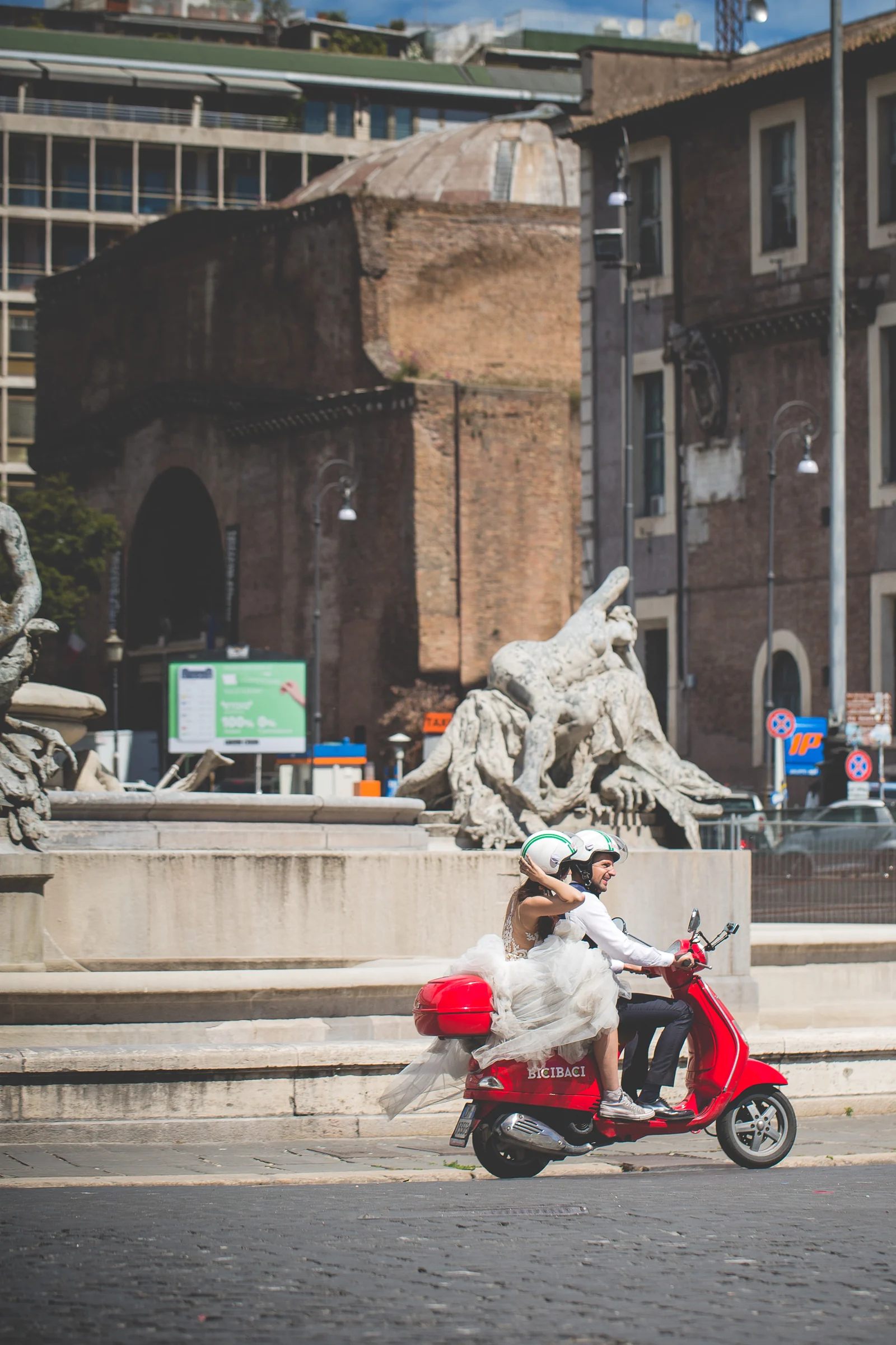 Los novios recorren Roma en una Vespa roja junto a una fuente monumental durante una sesión postboda en Calles de Roma con Vespa.
