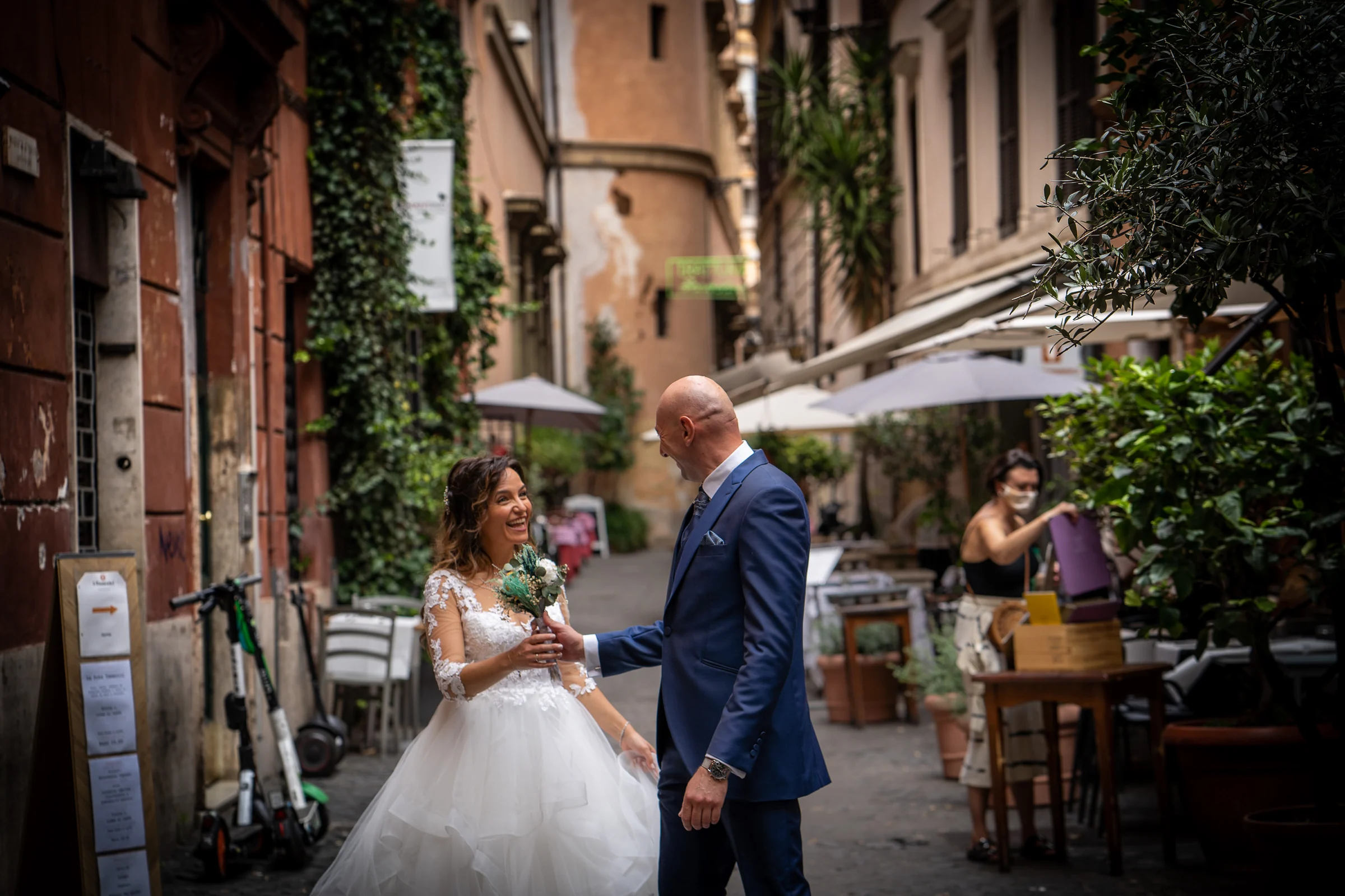 Marco y Esther ríen y se miran en una calle romana con terrazas y fachadas históricas durante su sesión postboda.