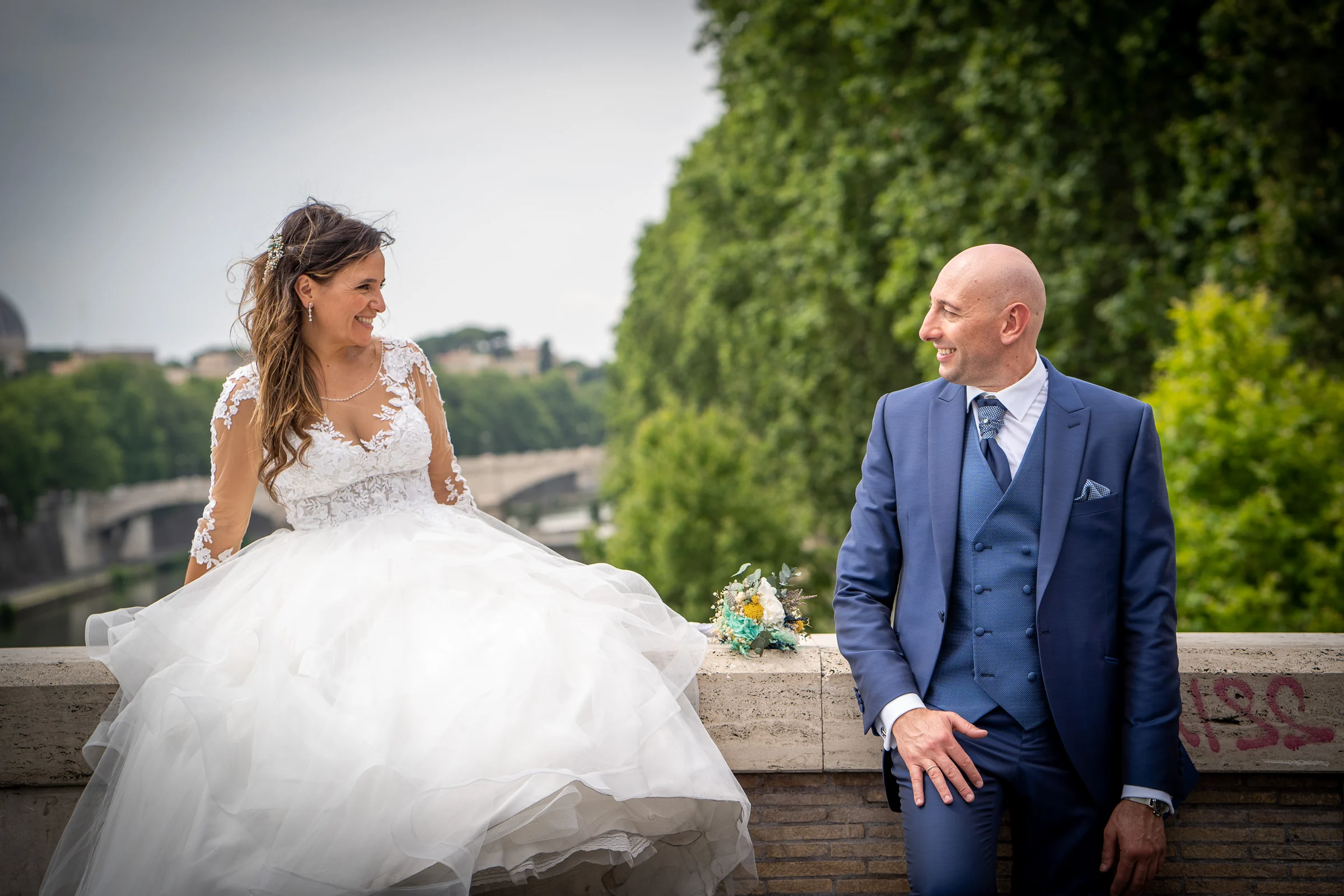 Marco y Esther se miran sentados junto al río Tíber con el paisaje verde de Roma al fondo durante su sesión postboda.