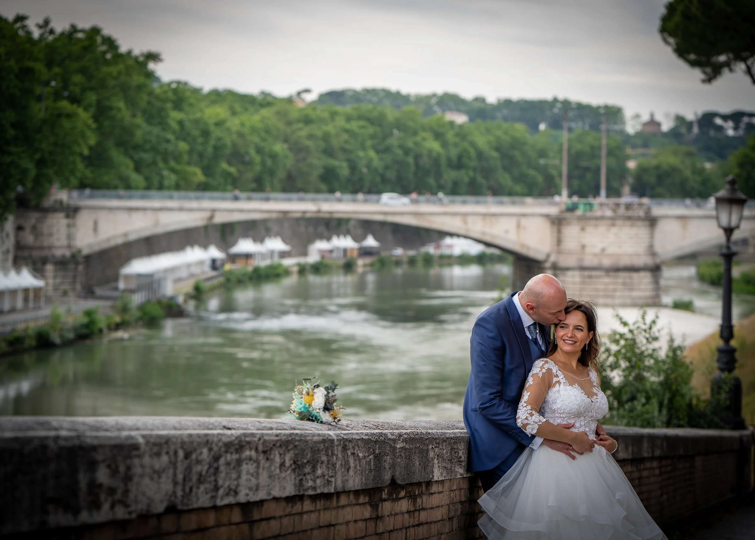 Marco abraza a Esther junto a una balaustrada con el río Tíber y un puente de Roma desenfocados al fondo.
