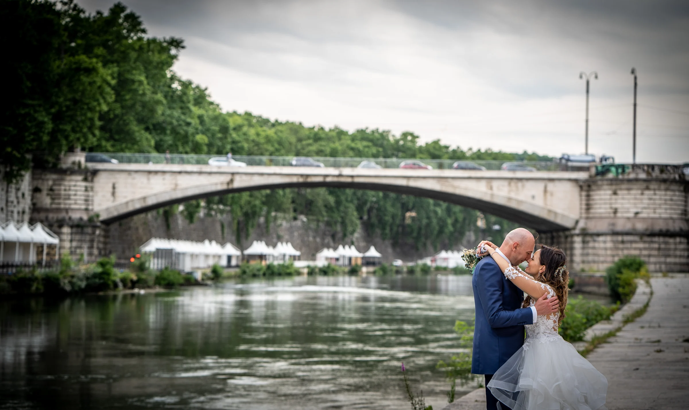 Marco y Esther se besan junto al río Tíber con el puente y la vegetación romana como escenario natural.