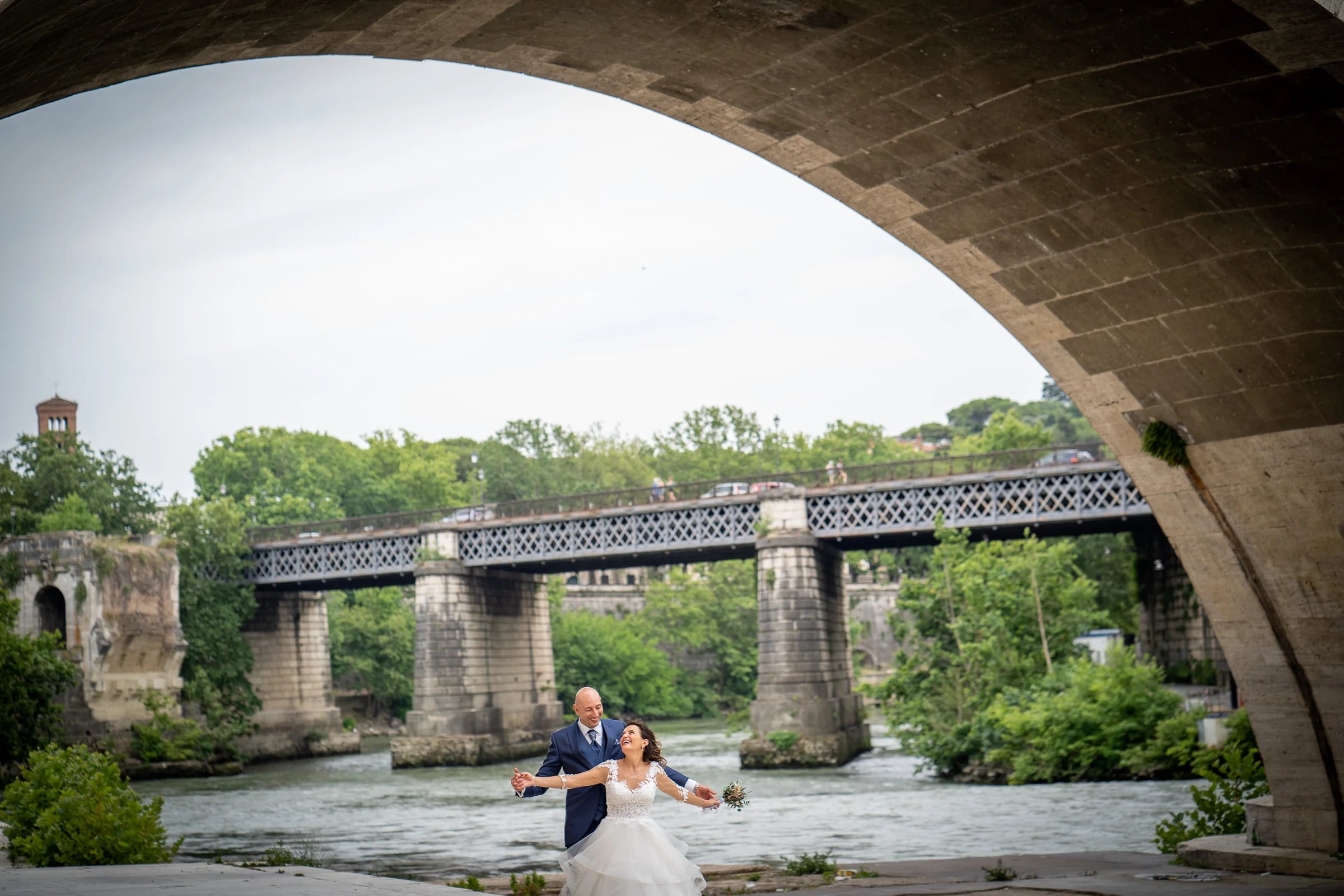 Marco y Esther se sientan juntos bajo un arco monumental junto al río Tíber durante su postboda en Roma.