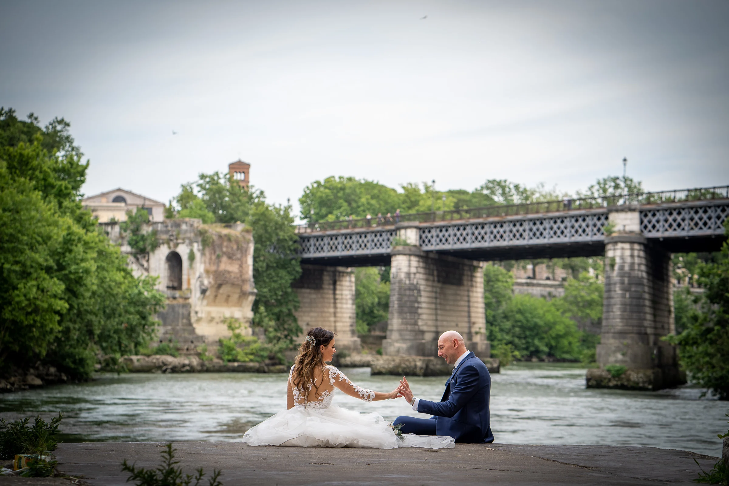 Marco y Esther se sientan frente al río Tíber y un puente ferroviario, en una escena serena y natural.