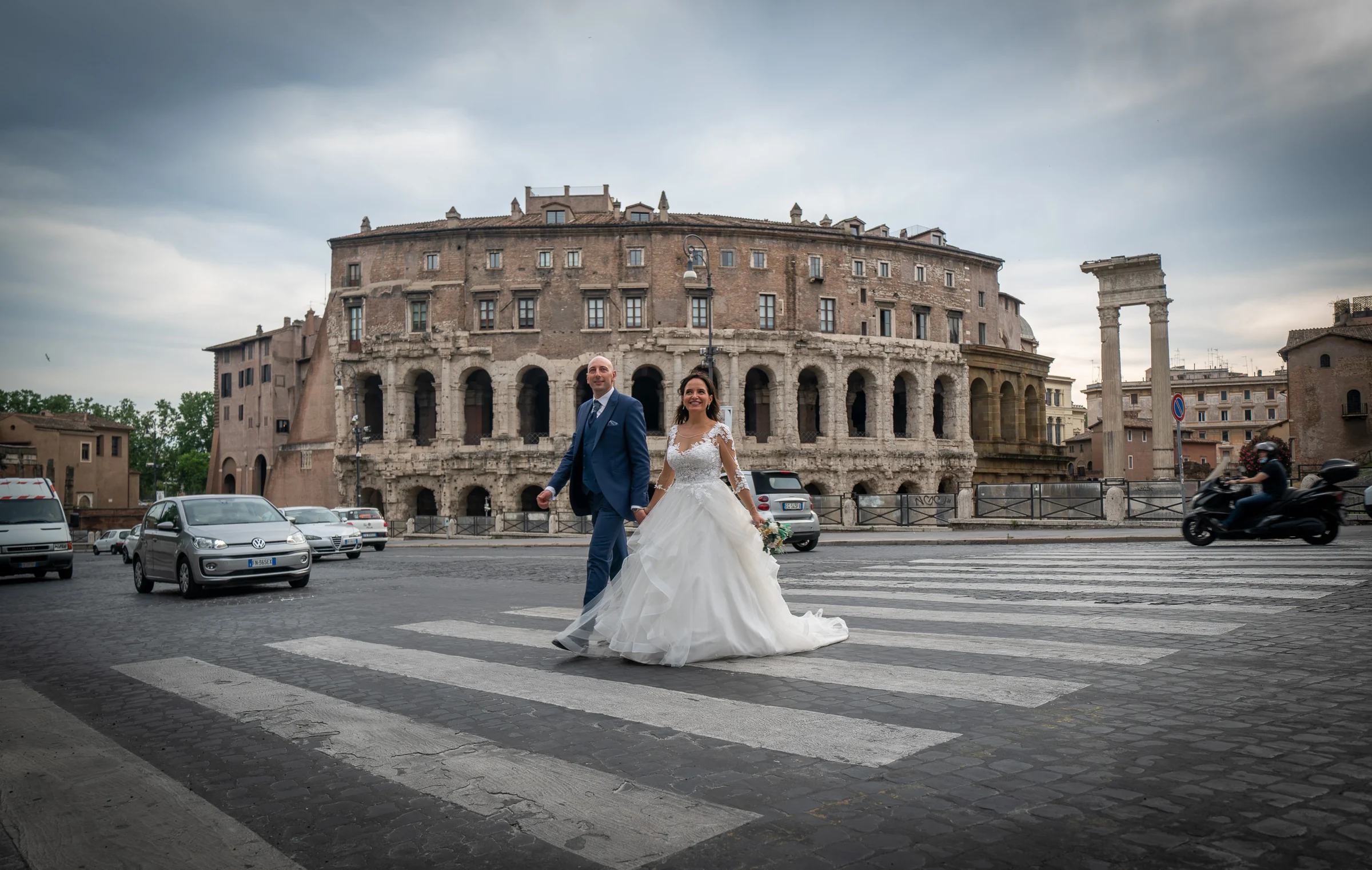 Marco y Esther cruzan una avenida frente a arquitectura romana antigua durante su reportaje de postboda en la ciudad.