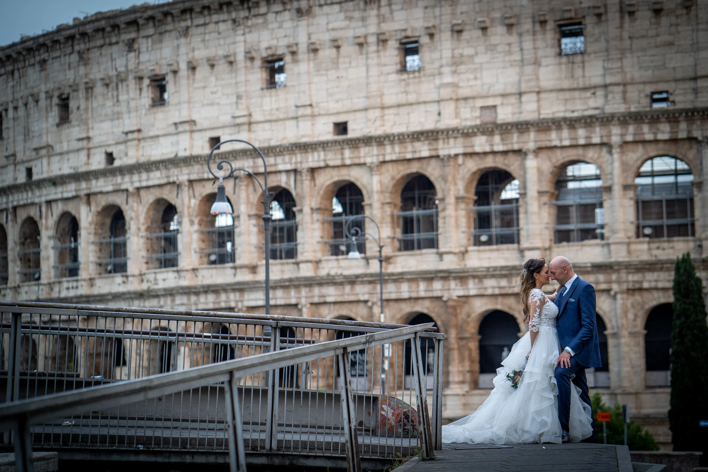 Marco y Esther posan en una pasarela elevada con la fachada del Coliseo ocupando gran parte de la escena.