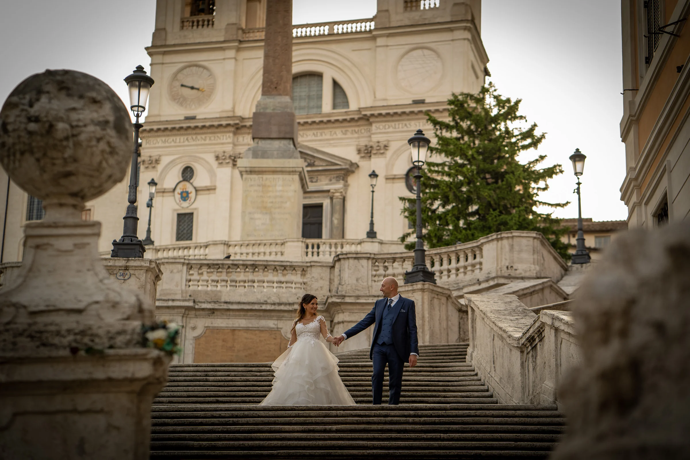 Marco y Esther bajan de la mano por una escalinata monumental de Roma con arquitectura clásica al fondo.