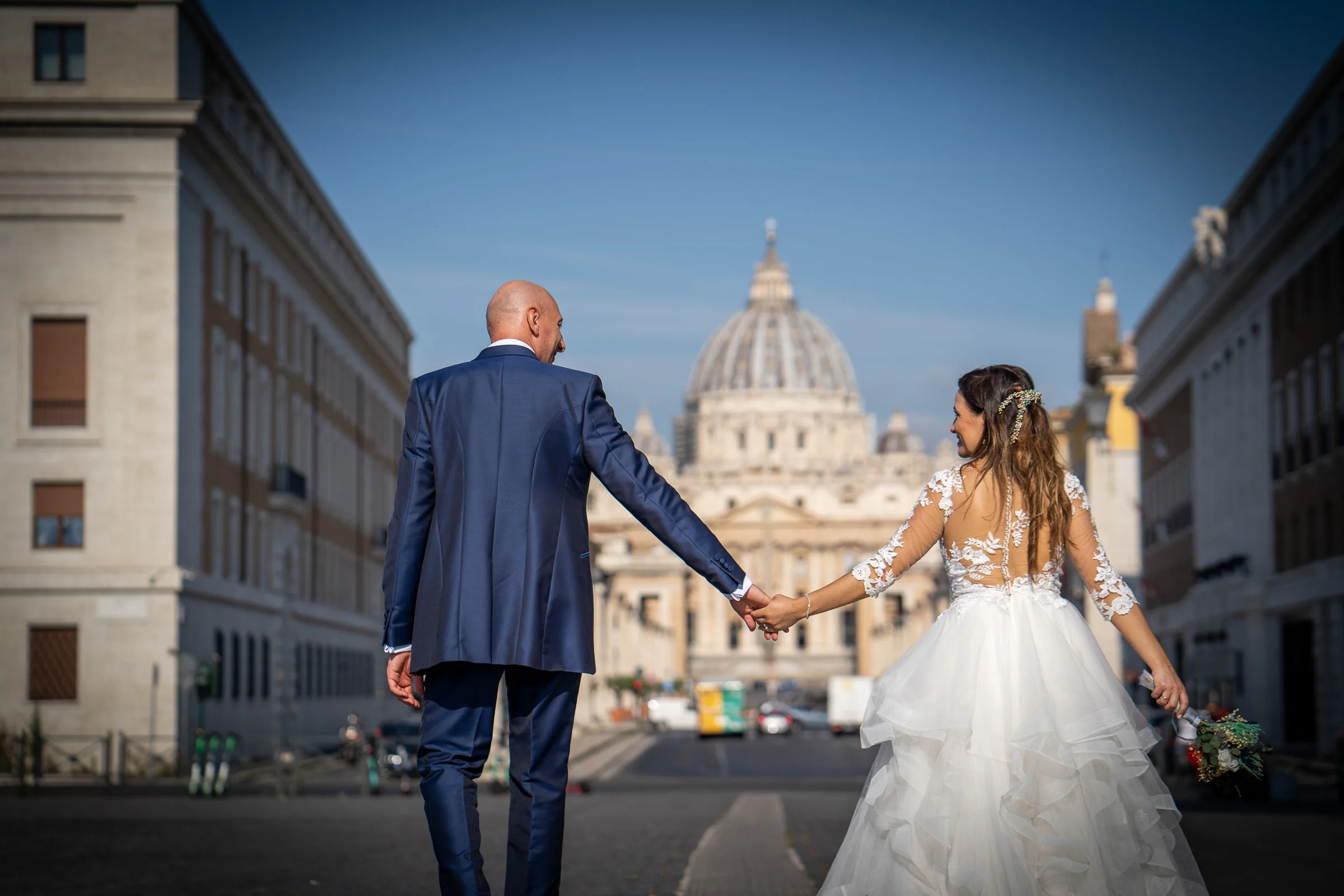 Marco y Esther caminan de la mano hacia la Basílica de San Pedro por una gran avenida de Roma.