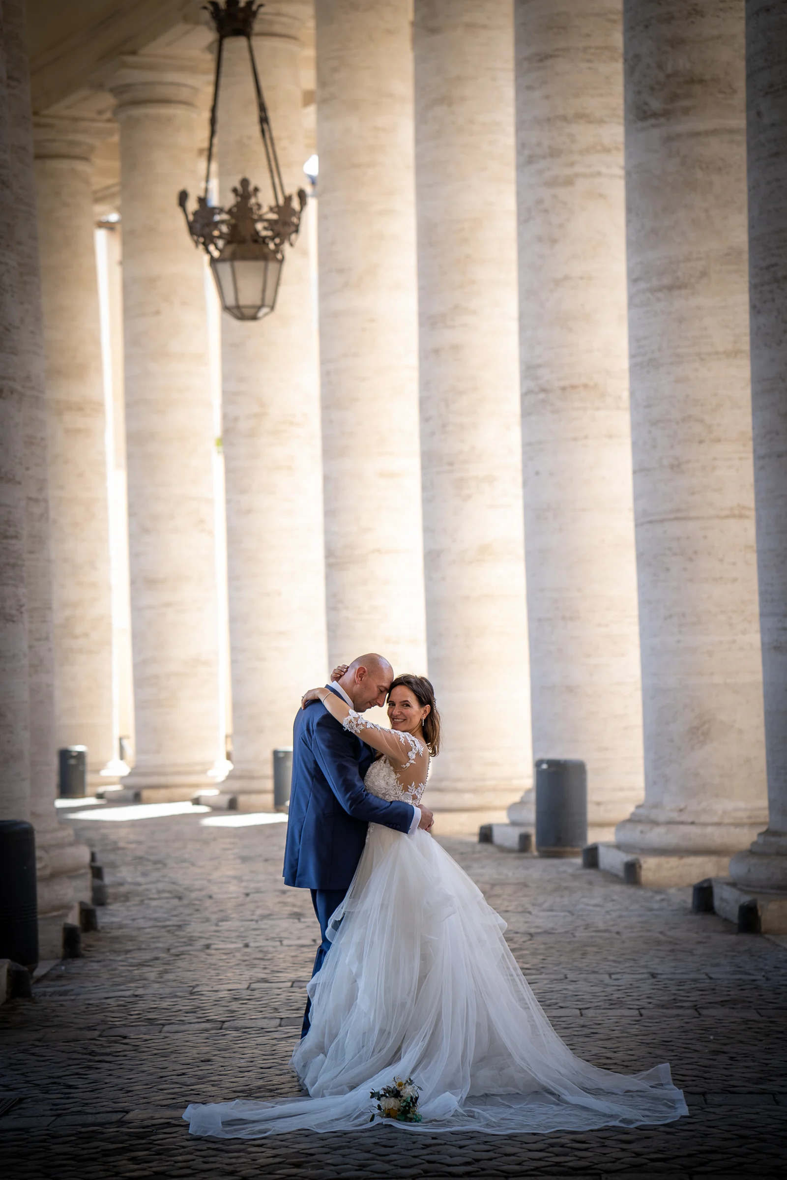 Marco abraza a Esther entre las columnas de la Plaza de San Pedro en un retrato vertical elegante.