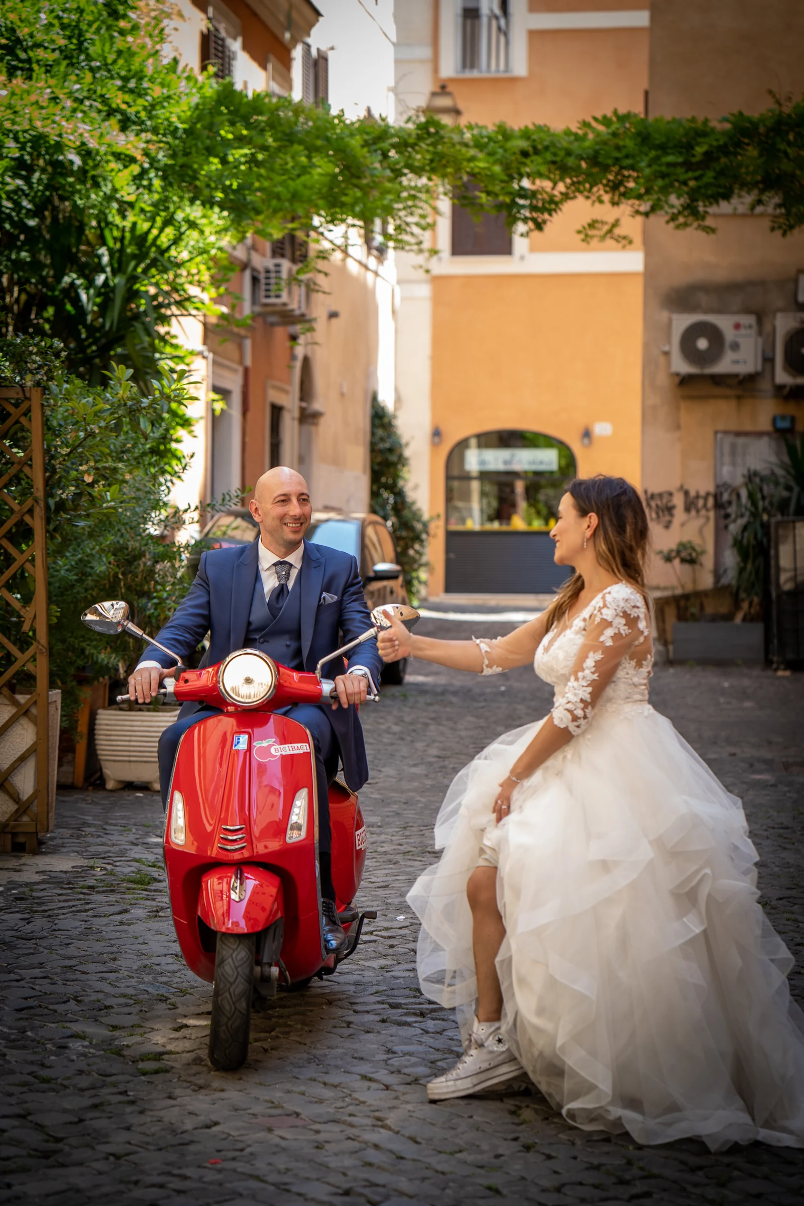 Marco posa sobre una Vespa roja mientras Esther le hace un gesto divertido en una calle romana.