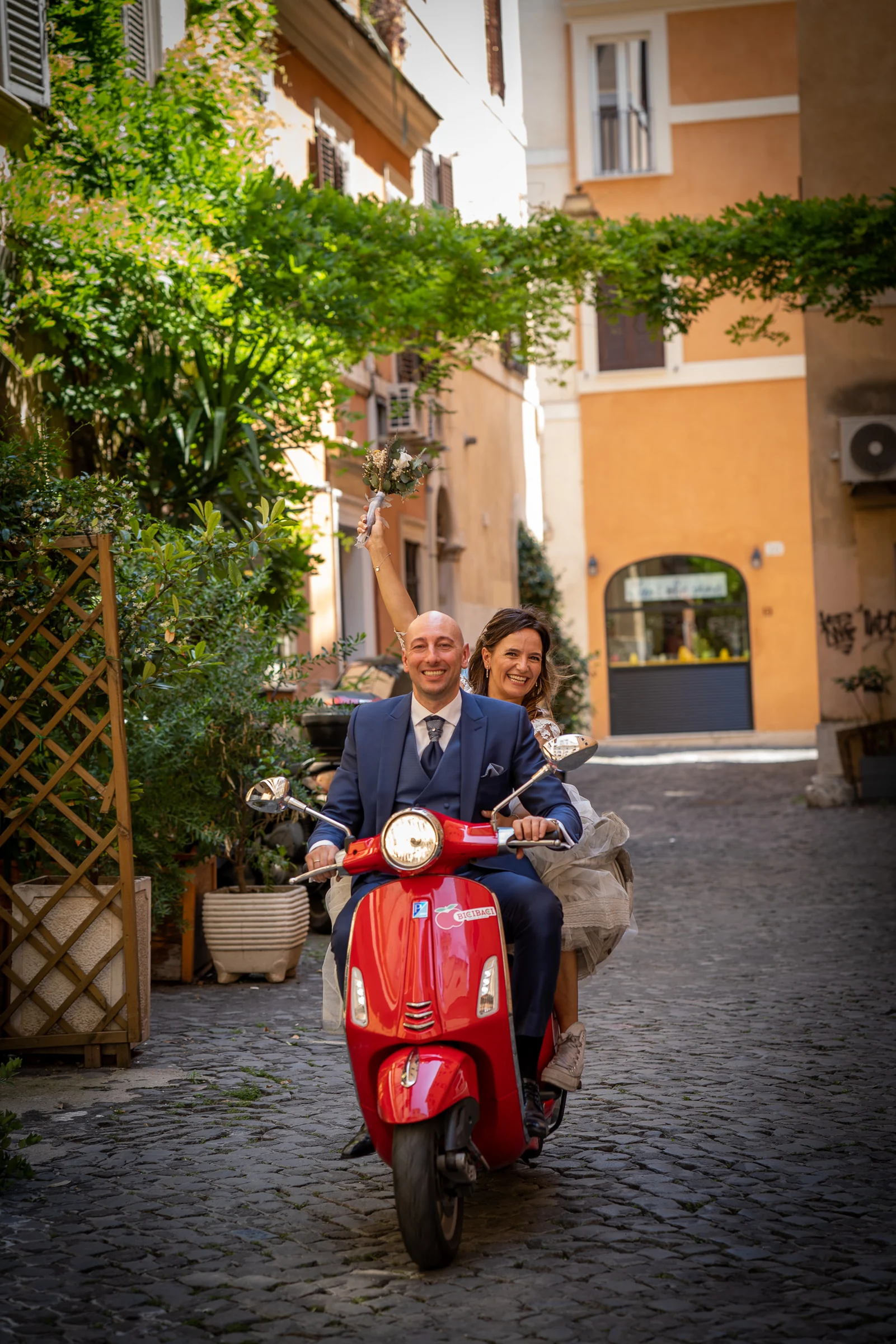 Marco y Esther sonríen juntos sobre una Vespa roja en una calle adoquinada del centro histórico de Roma.