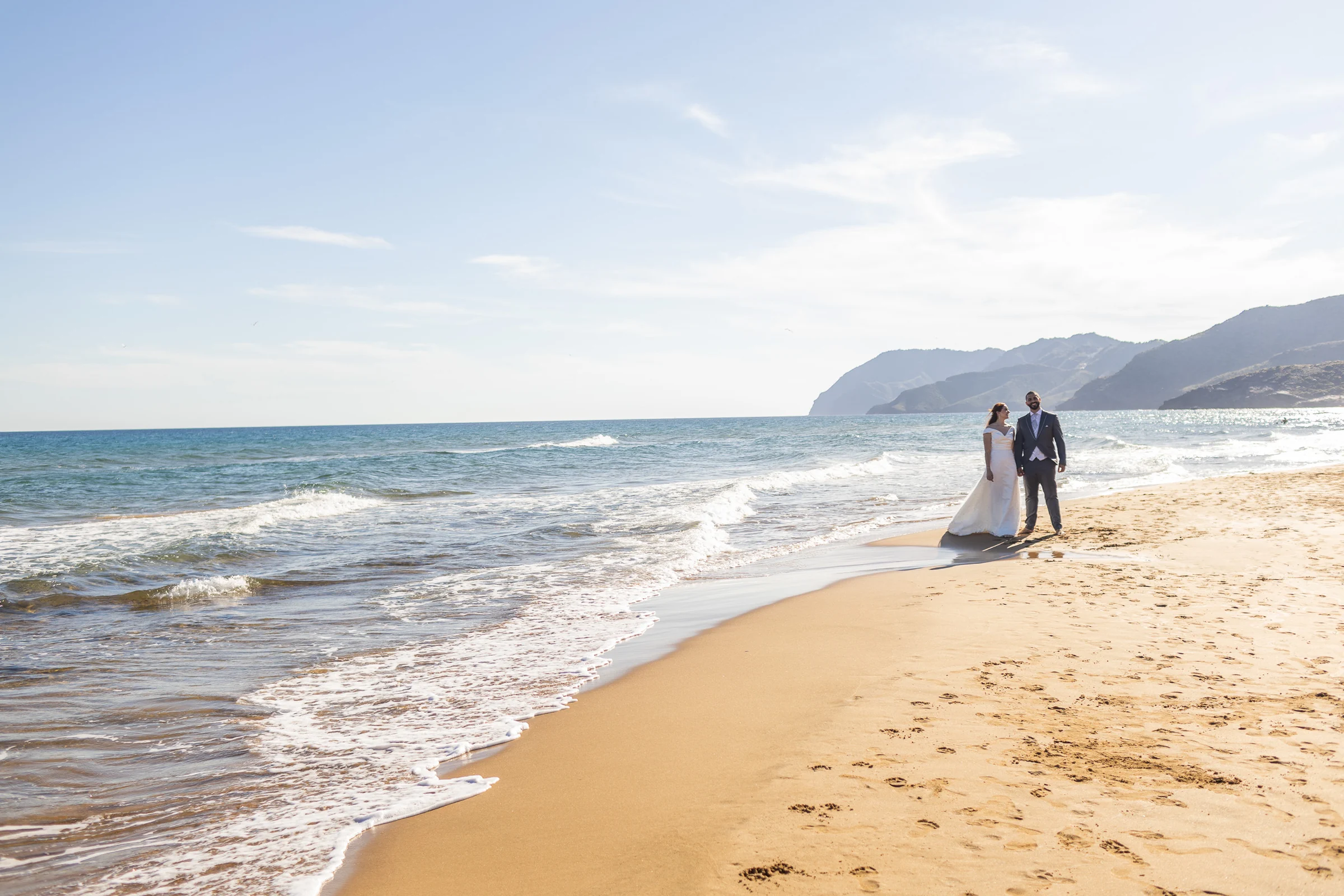 Noemí y Fran pasean por una playa amplia con el mar y las montañas del litoral de Cartagena al fondo.
