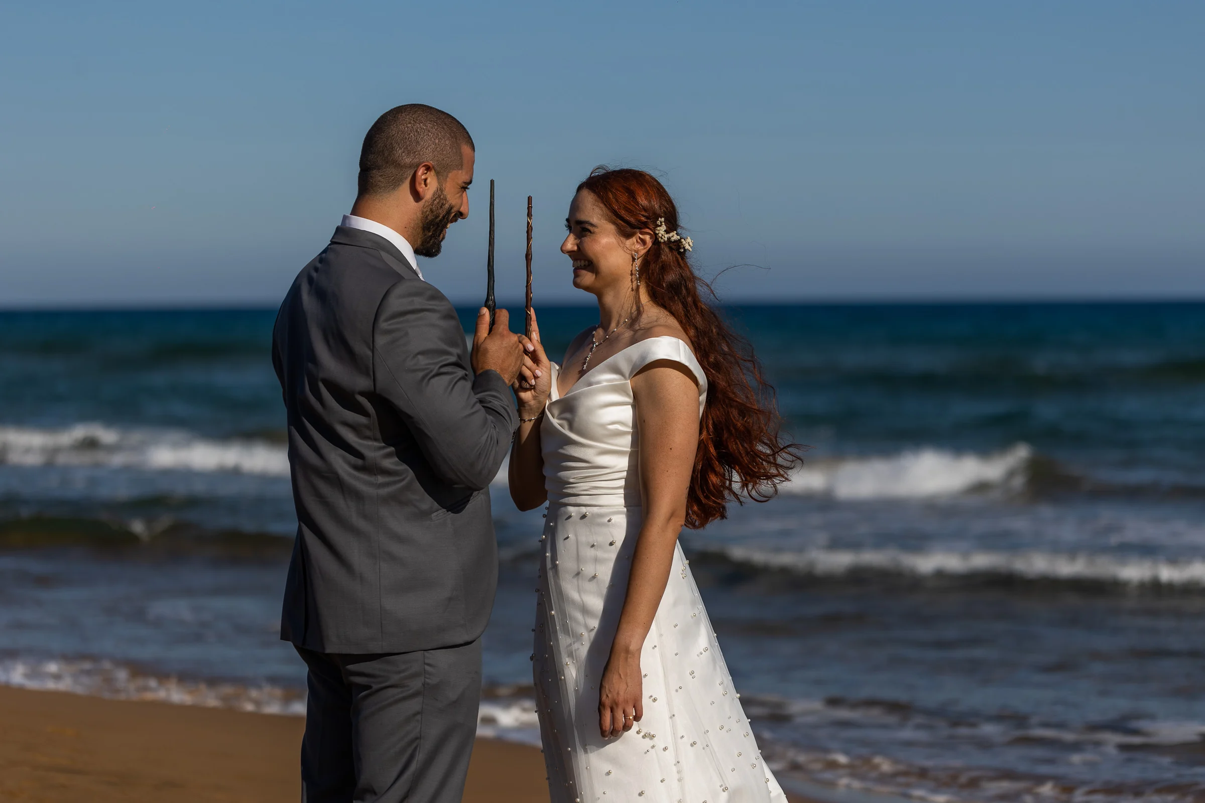Noemí y Fran sonríen frente a frente sosteniendo sus varitas en la playa durante su postboda en Cartagena.