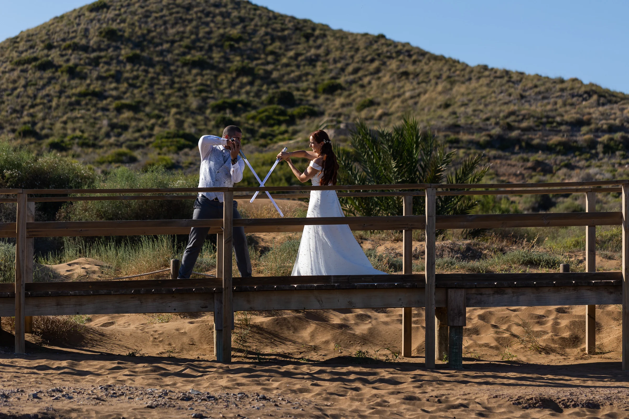Noemí y Fran juegan con sus varitas sobre una pasarela de madera entre dunas durante su postboda en Cartagena.