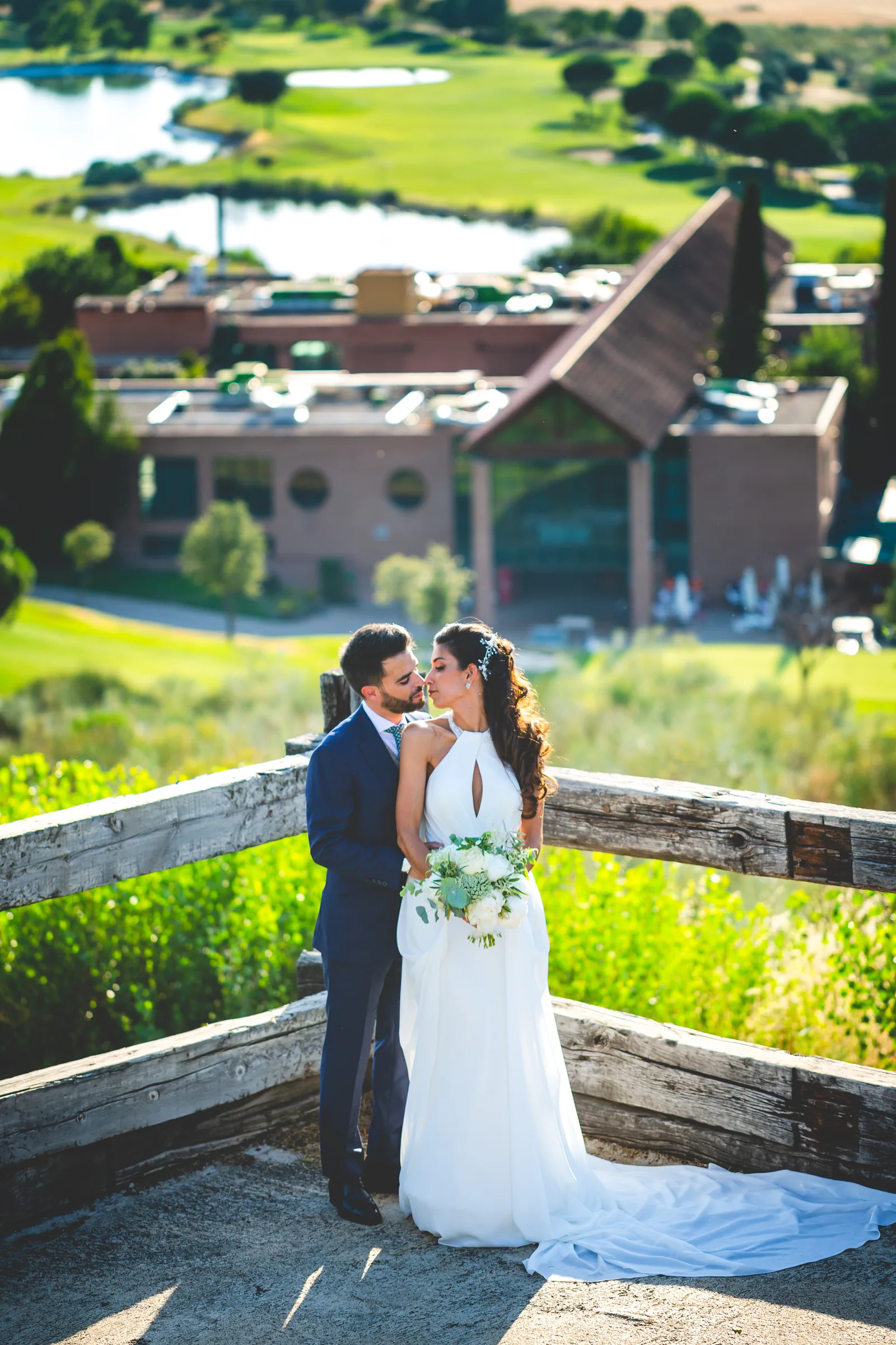 Los novios en un mirador de madera con el hotel y el campo de golf al fondo en Retamares Experience.