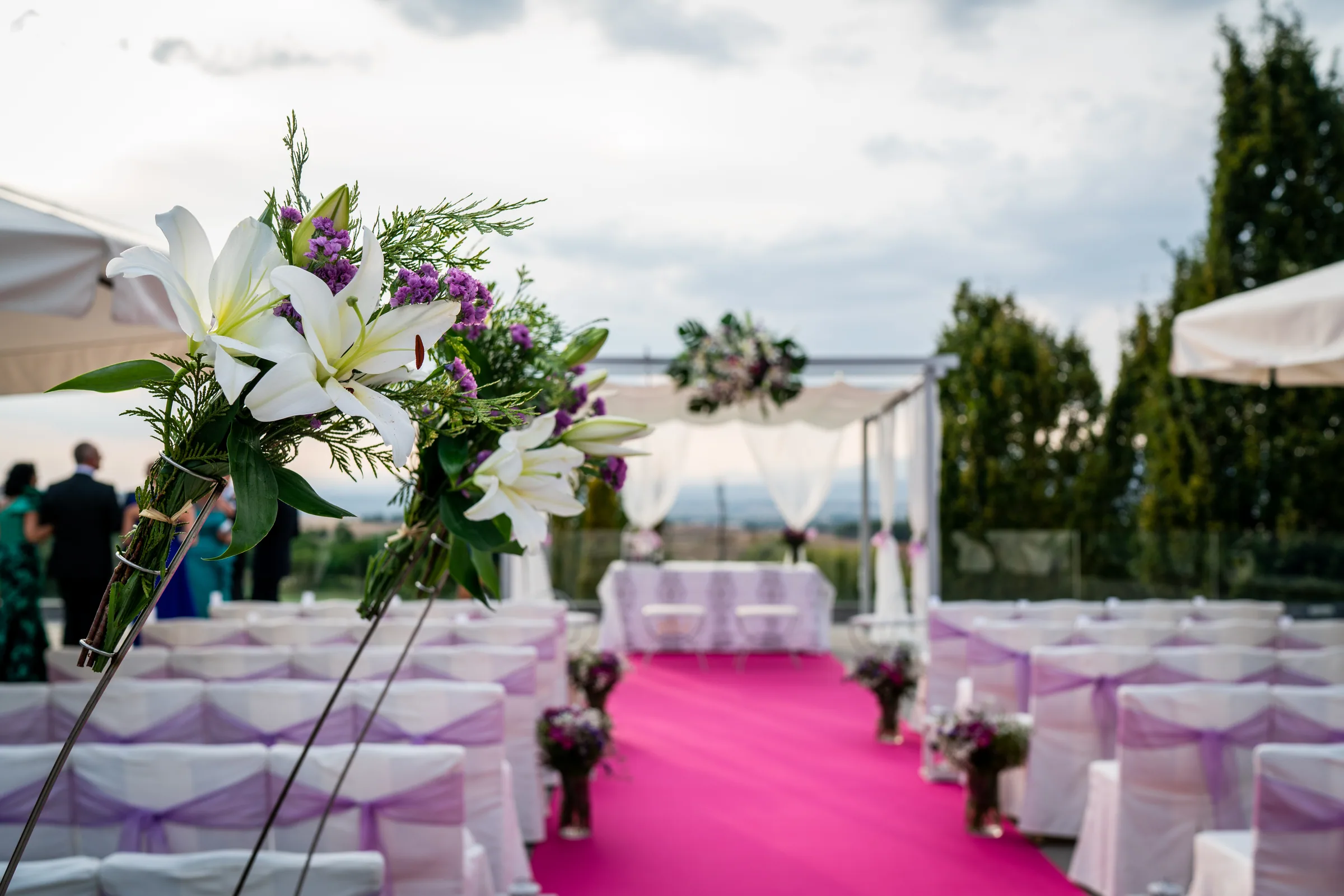 Pasillo fucsia entre sillas con lazos lilas y grandes arreglos de lirios blancos en la terraza de la ceremonia en Retamares Experience.