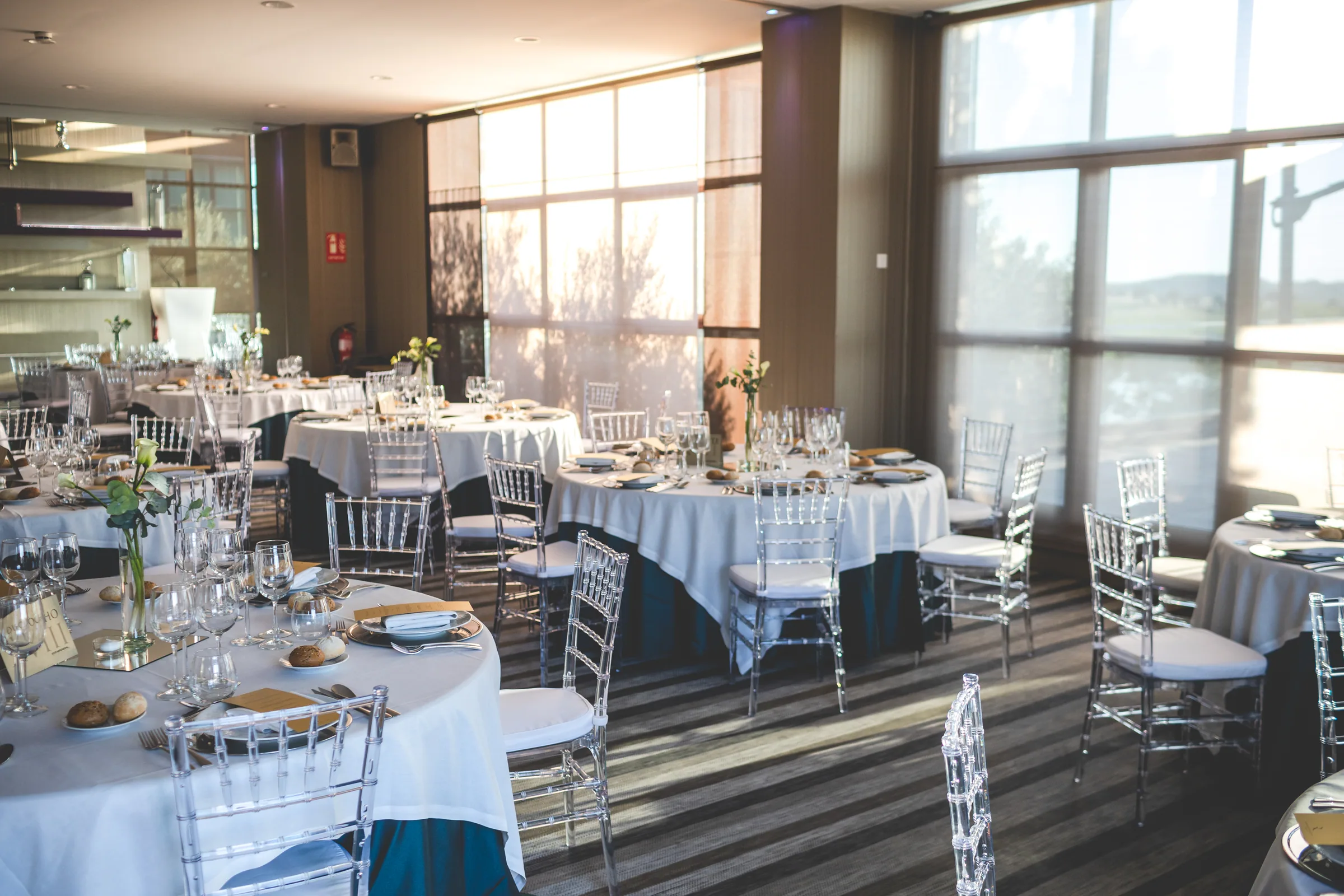 Salón del banquete montado con mesas redondas, sillas transparentes y grandes ventanales en Retamares Experience.
