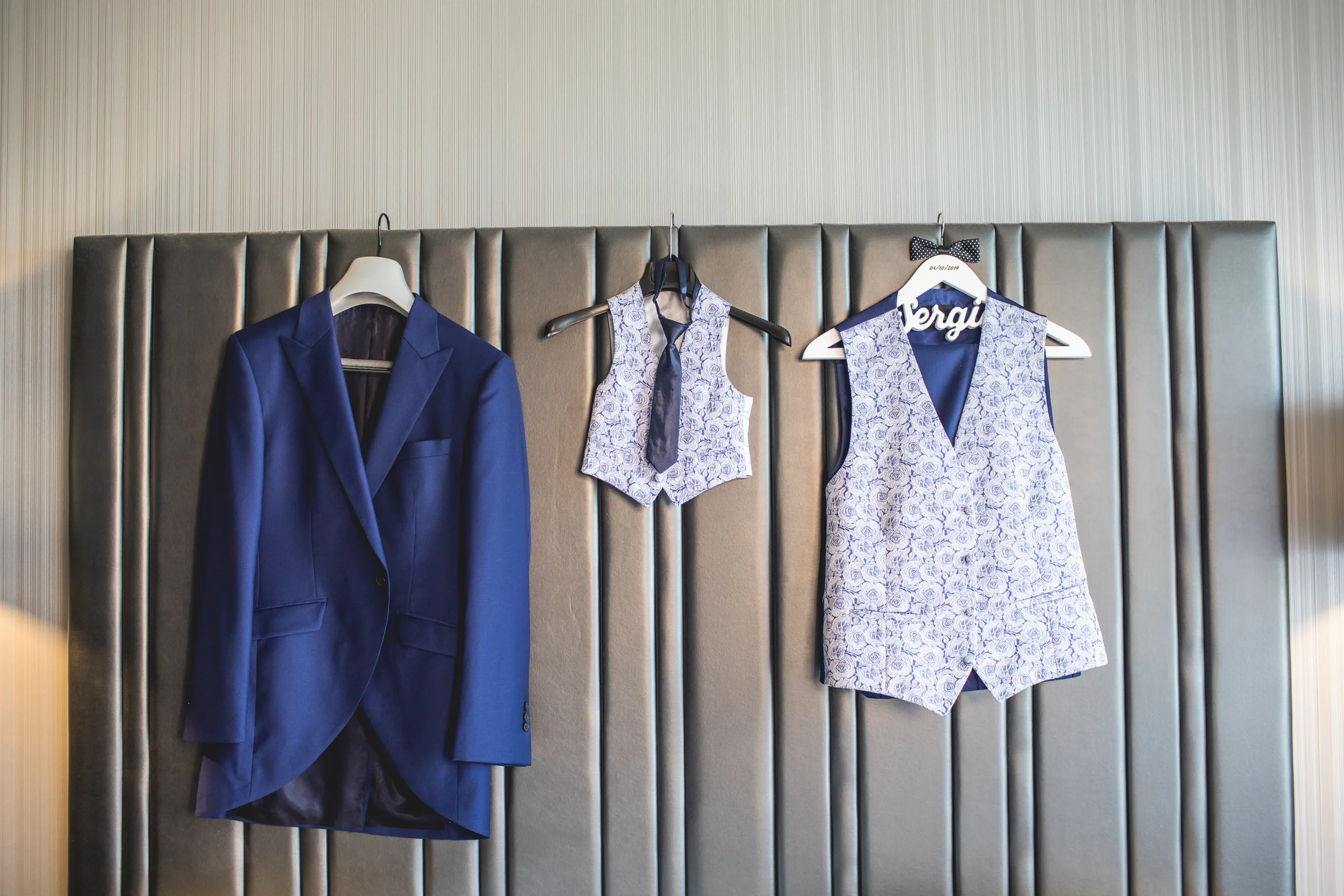 Traje azul del novio, chaleco estampado y corbata colgados sobre un cabecero en una habitación del hotel en Retamares Experience.