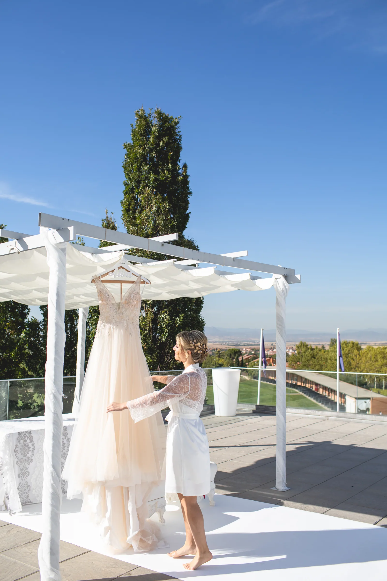 El vestido de novia colgado en una pérgola blanca de la terraza con el paisaje al fondo en Retamares Experience.