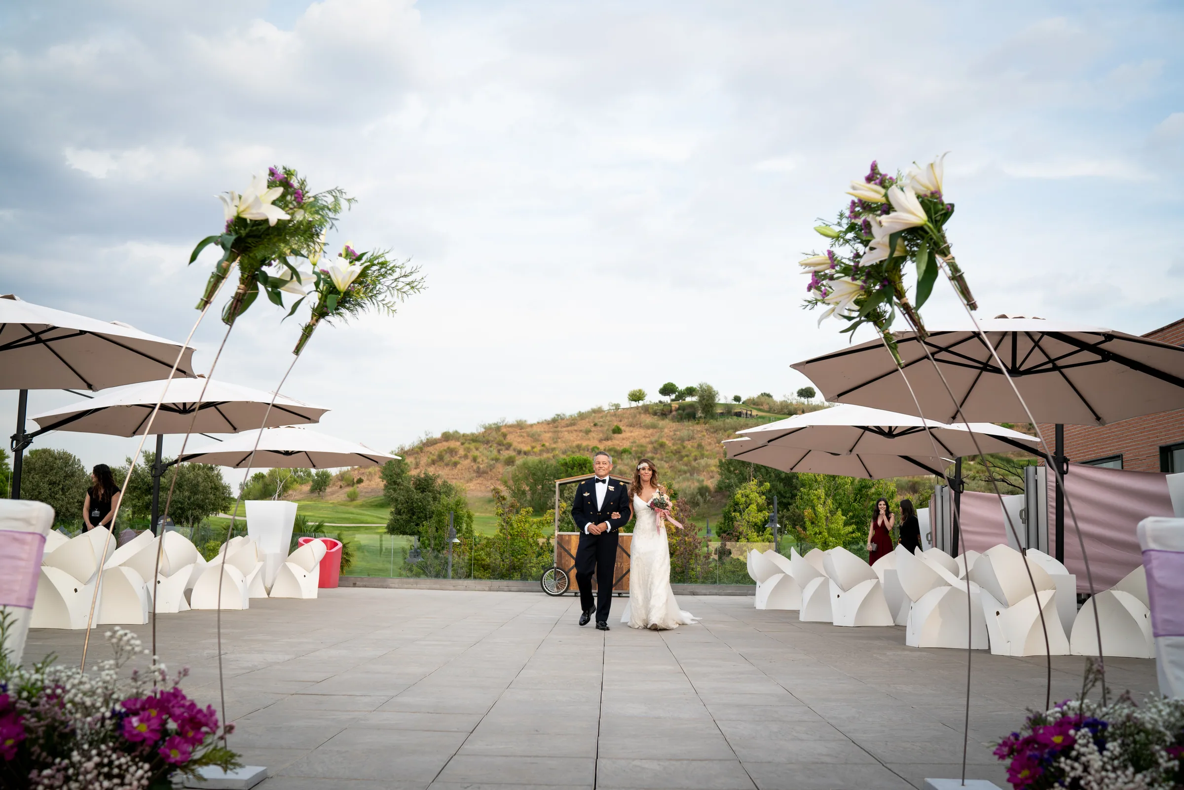 La novia camina por la terraza hacia la ceremonia entre sombrillas y arreglos florales en Retamares Experience.