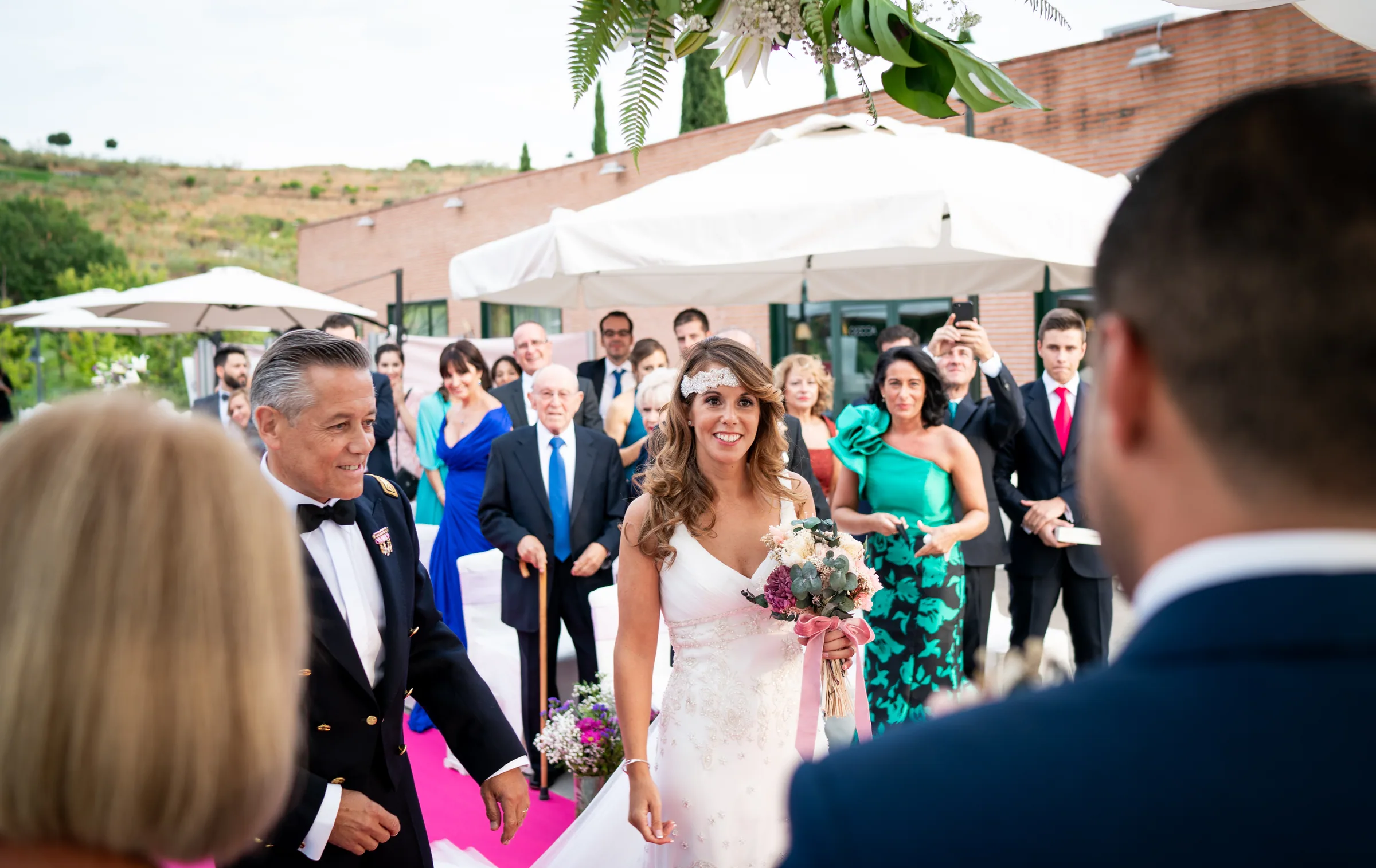 La novia entra del brazo de su padre por el pasillo fucsia rodeada de invitados en Retamares Experience.