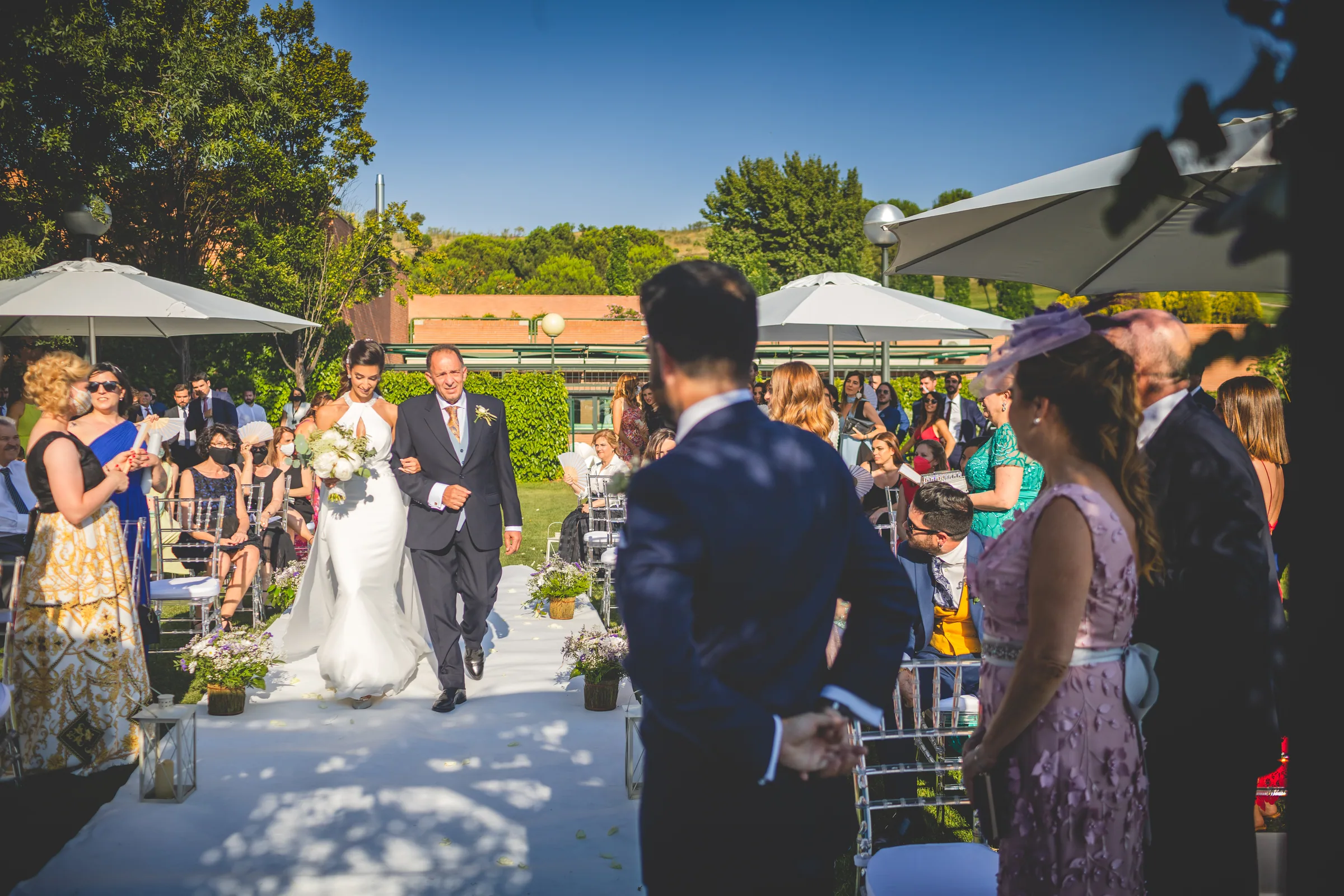 La novia entra a la ceremonia del brazo de su padre por el pasillo entre los invitados en Retamares Experience.
