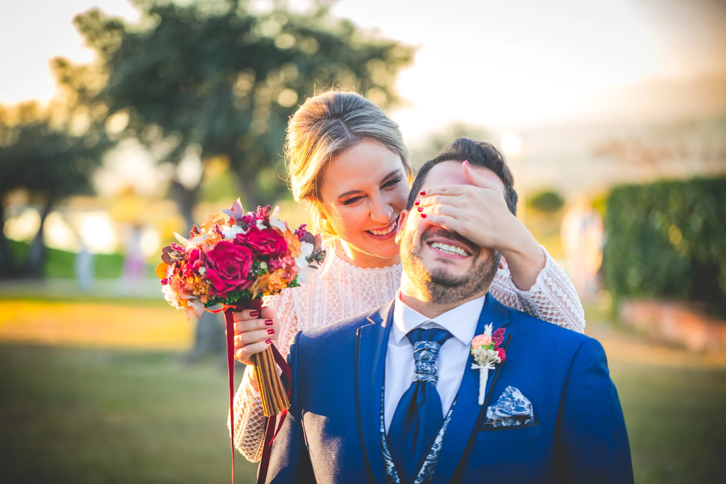 La novia tapa los ojos del novio con una sonrisa antes de verse, con un ramo rojo en Retamares Experience.