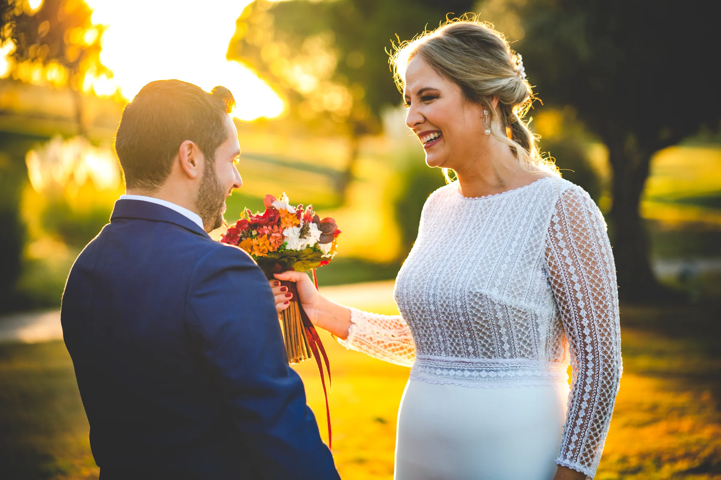 Los novios se miran sonriendo con el ramo rojo y la luz dorada en Retamares Experience.