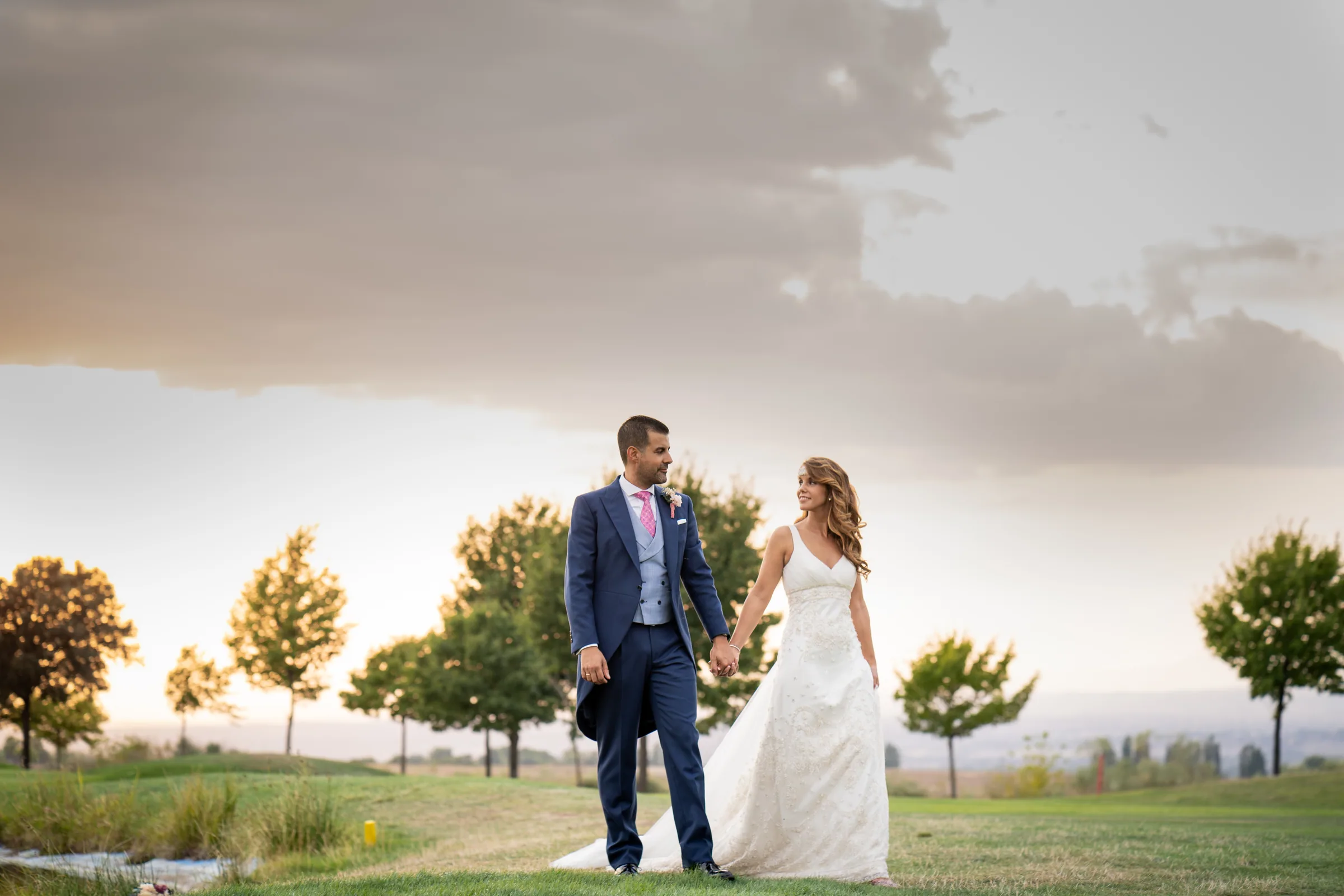 Los novios pasean de la mano por el césped junto al lago bajo un cielo con nubes en Retamares Experience.