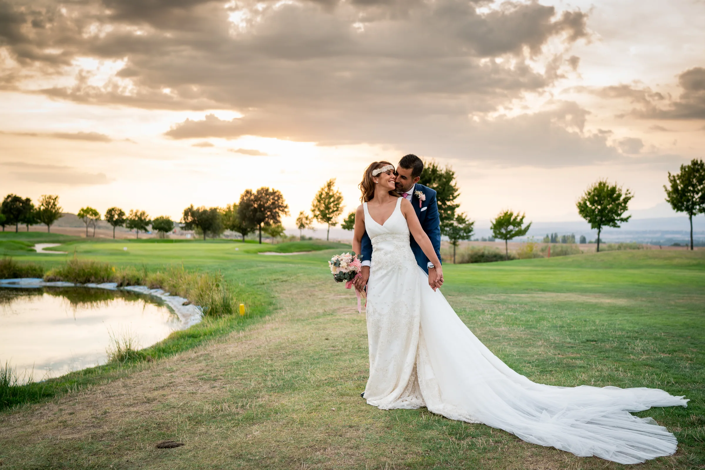 La novia abraza al novio con la cola del vestido extendida sobre el césped junto al lago en Retamares Experience.