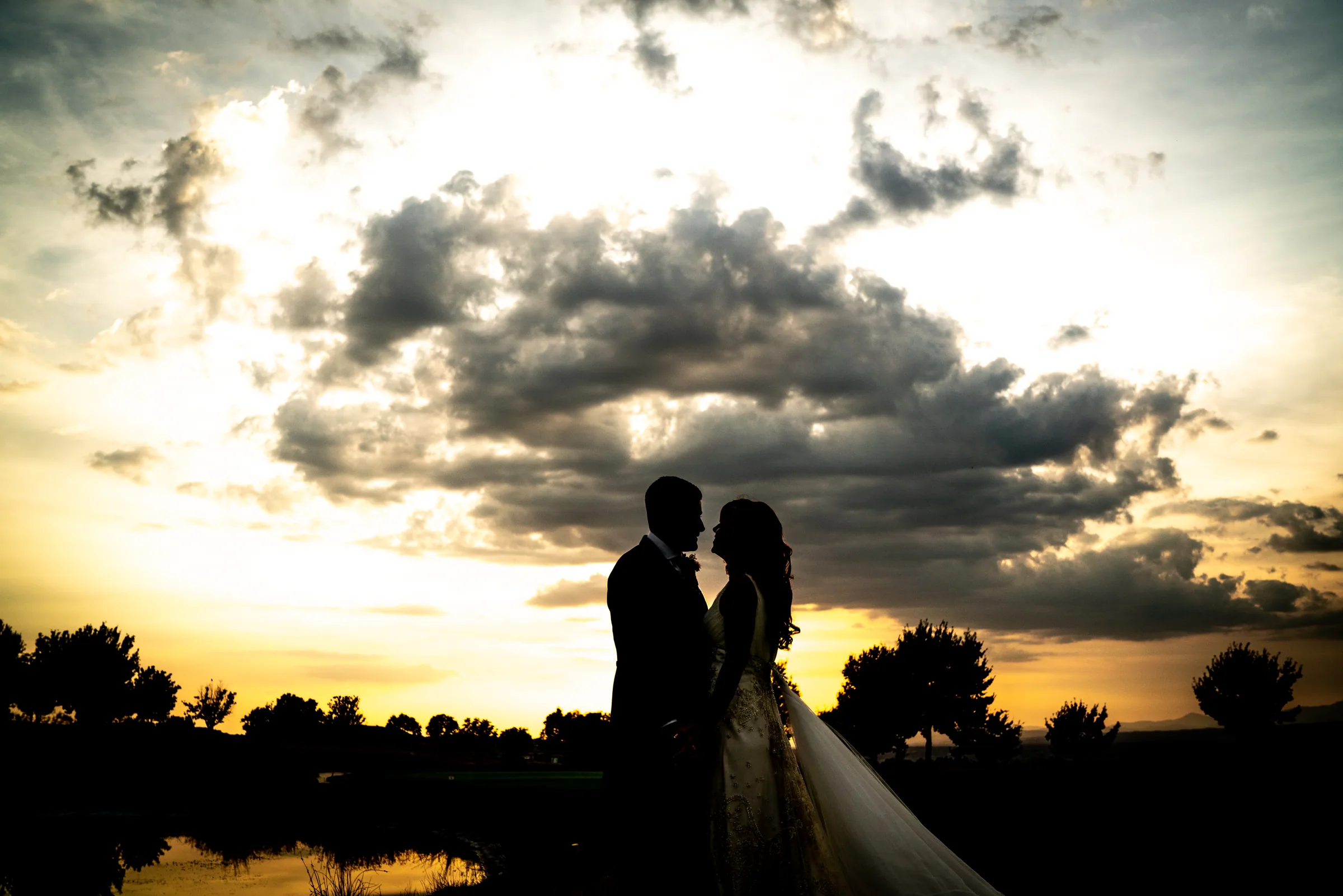 Silueta de los novios abrazados bajo un cielo dramático al atardecer junto al lago en Retamares Experience.