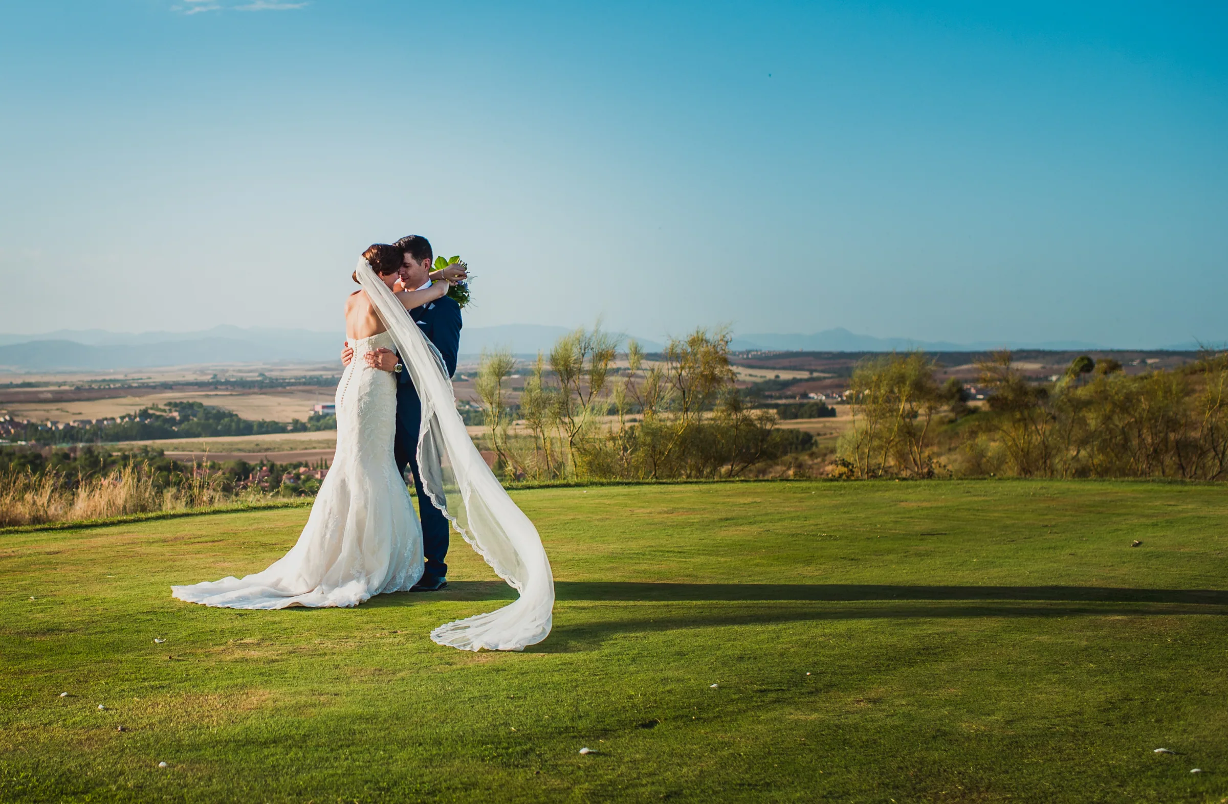 Los novios abrazados sobre el césped con el velo al viento y el paisaje al fondo en Retamares Experience.