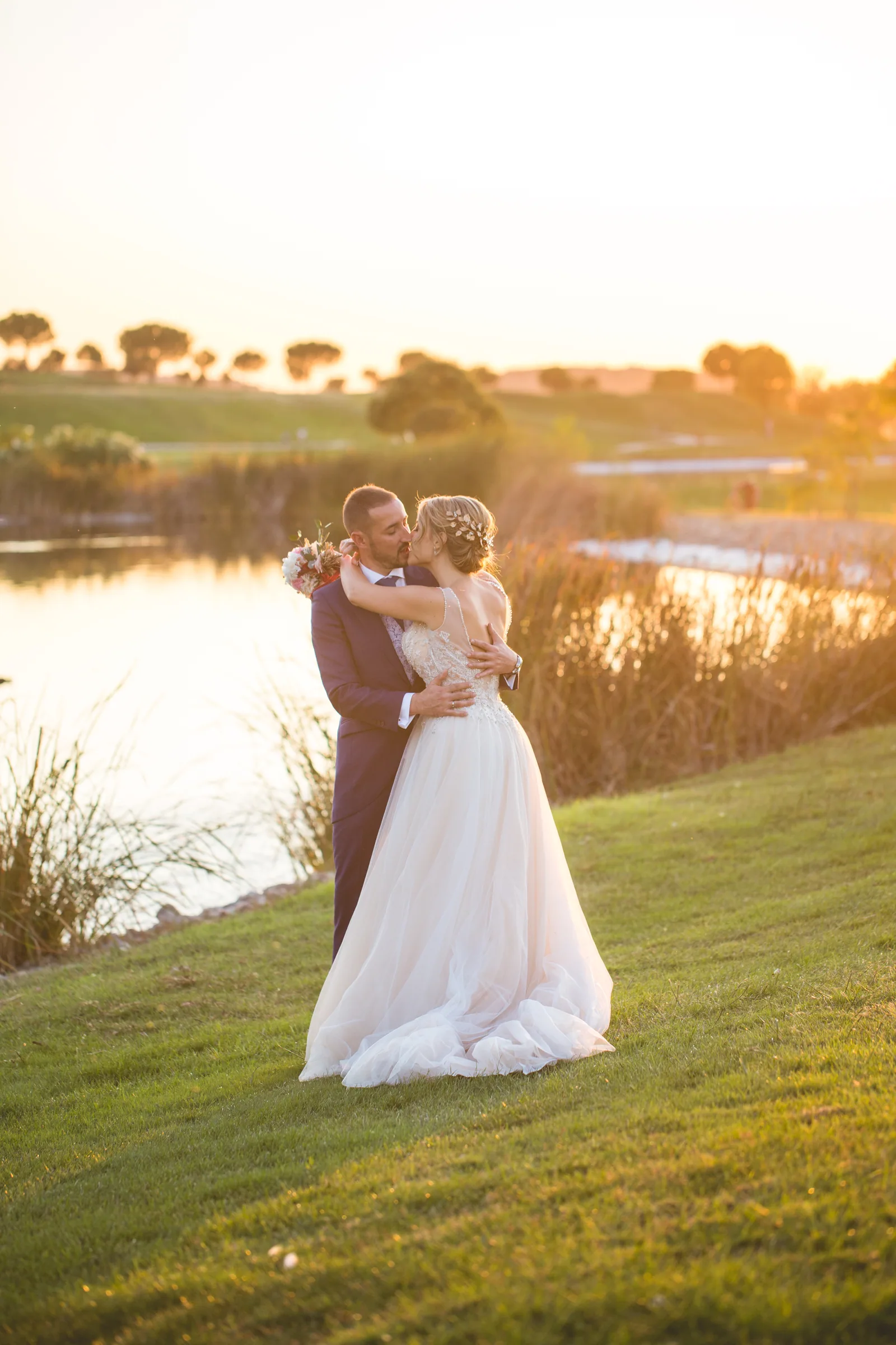 Los novios se besan sobre el césped junto al lago al atardecer en Retamares Experience.