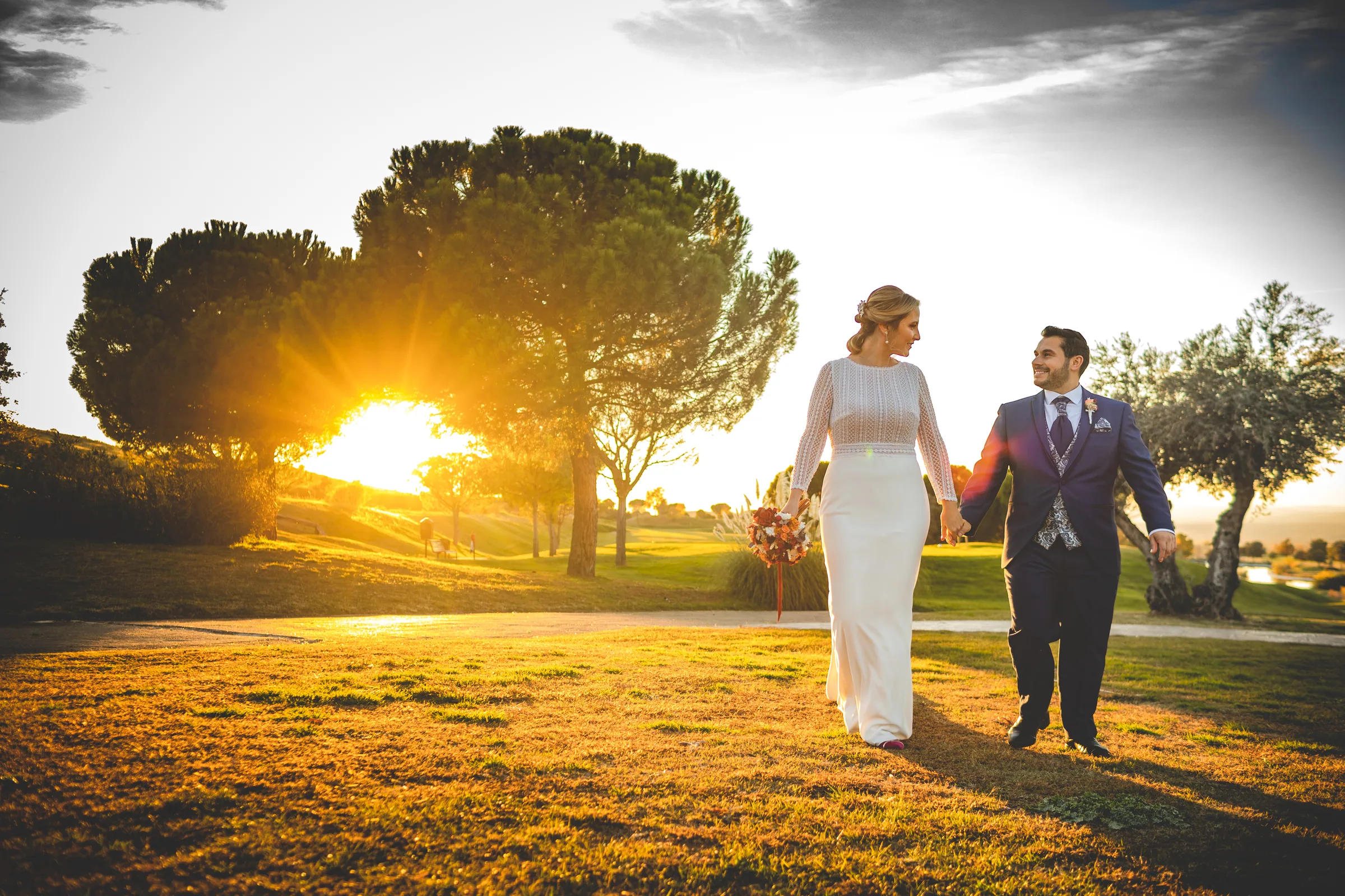 Los novios caminan de la mano hacia el sol poniente entre pinos en Retamares Experience.