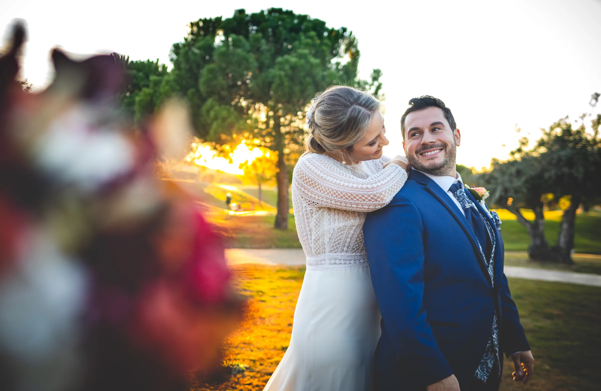 Los novios sonríen con flores desenfocadas en primer plano y el sol detrás en Retamares Experience.