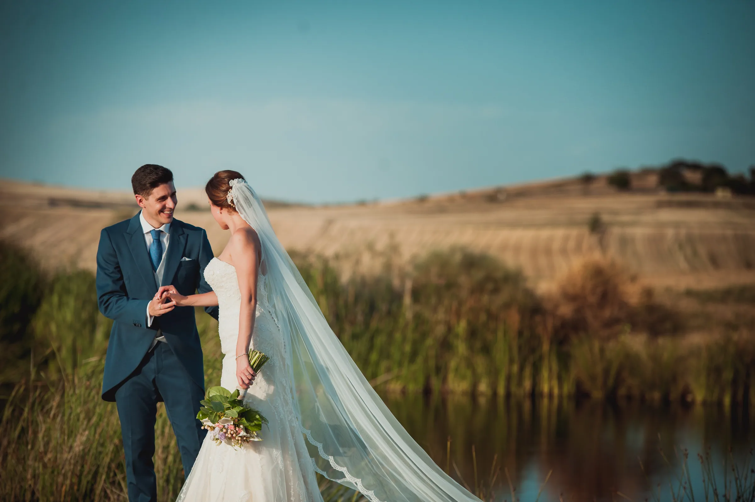 Los novios entre campos dorados con el velo al viento junto a un lago en Retamares Experience.